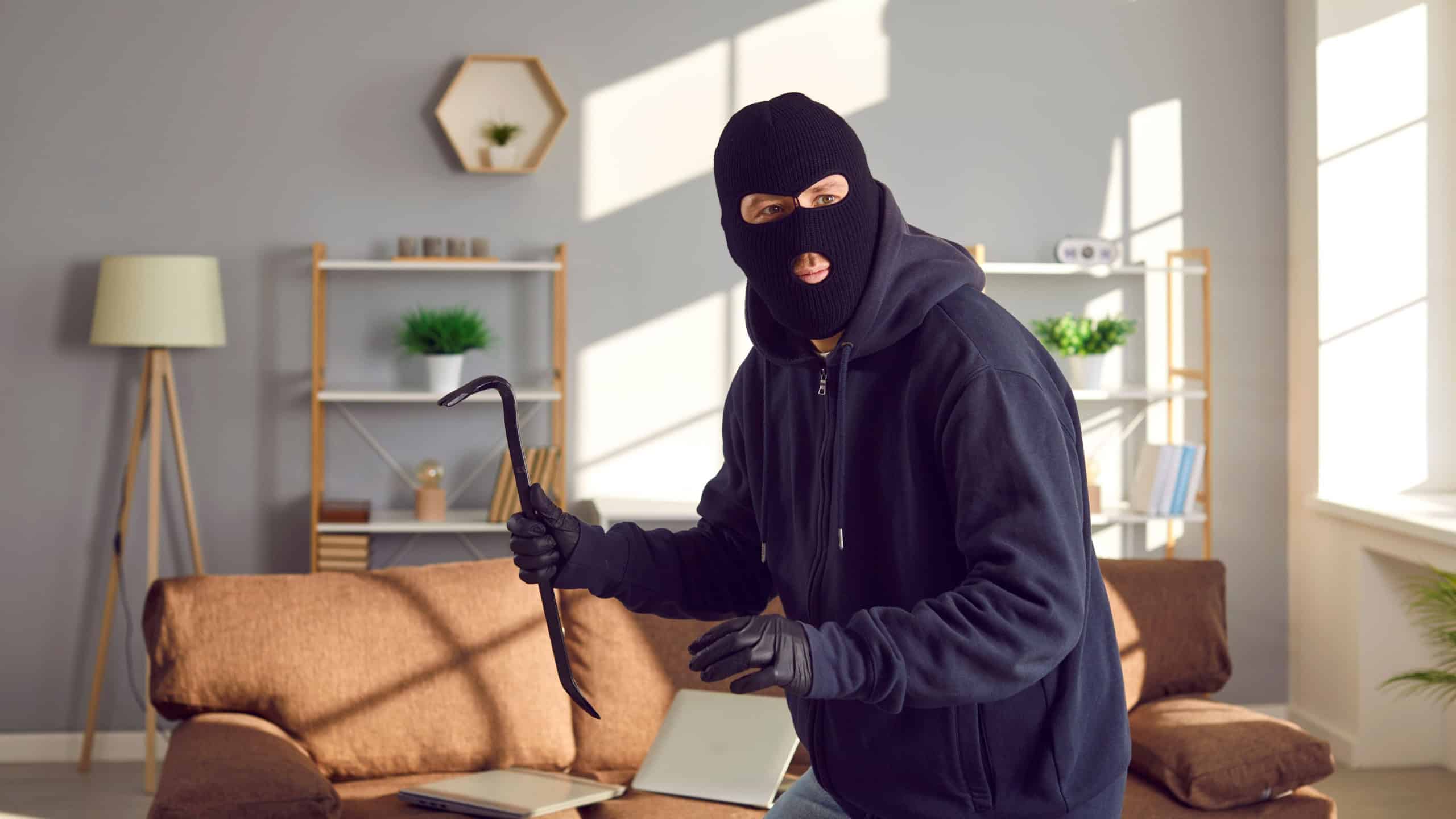 Burglar, robber, thief breaks into somebody's home. Man wearing black balaclava and gloves holding crowbar, walking on tiptoes and looking around for things he can take from this house or apartment