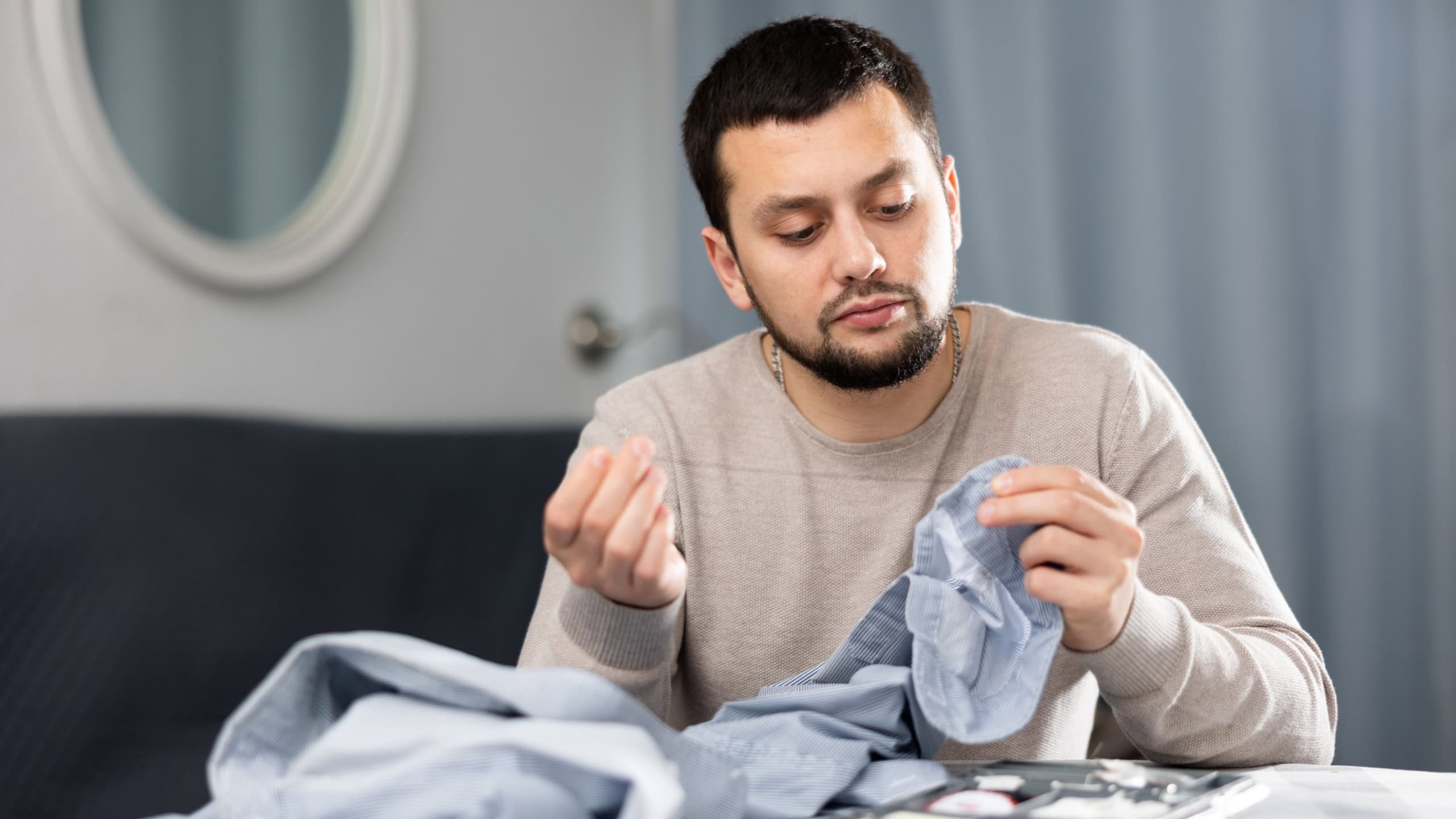 Young bearded man sitting at table in living room and sewing on buttons on sleeve of his shirt