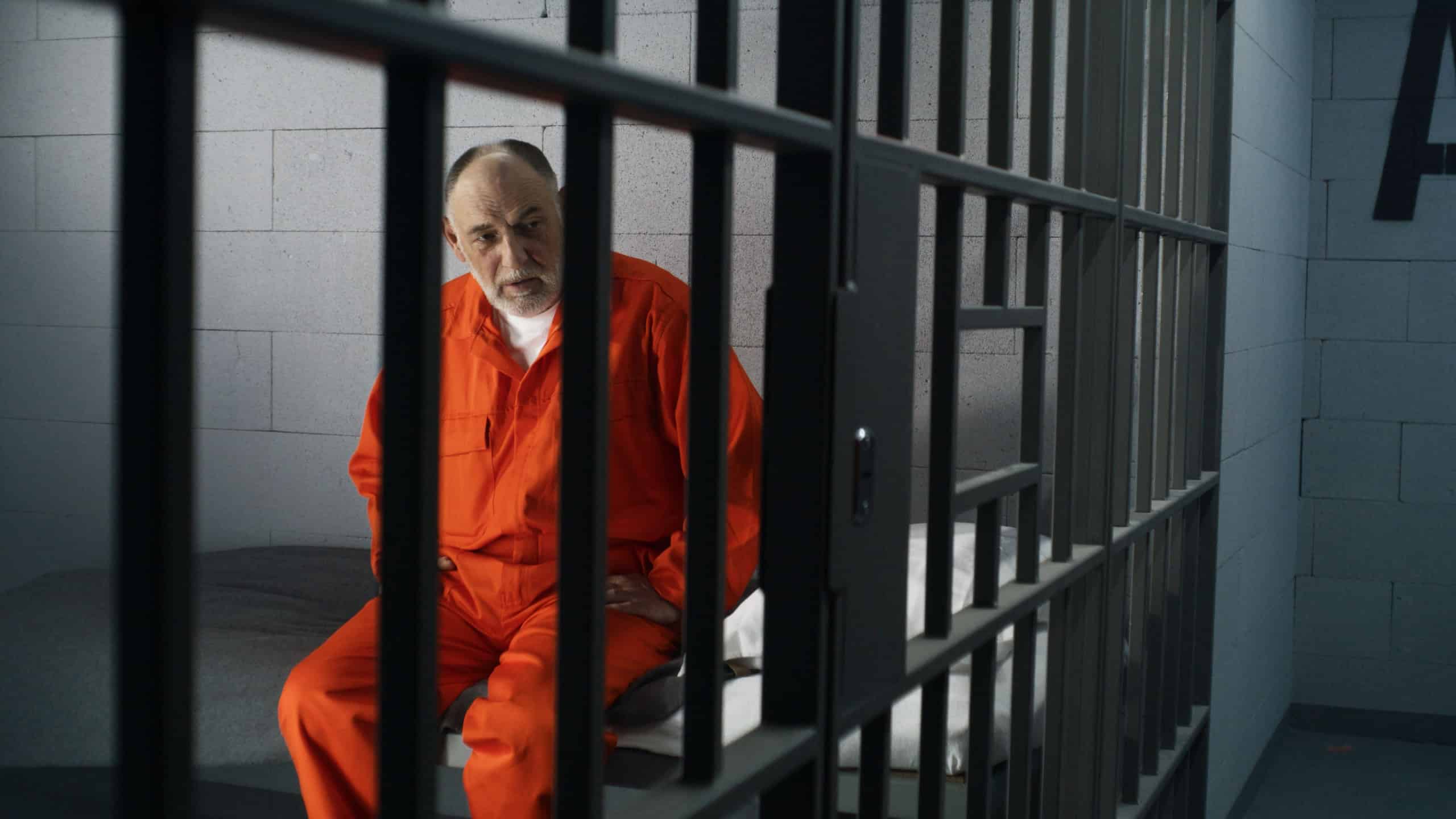 Elderly criminal in orange uniform sits on prison bed and thinks about freedom. Prisoner serves imprisonment term in jail cell. Guilty inmate in detention center or correctional facility.