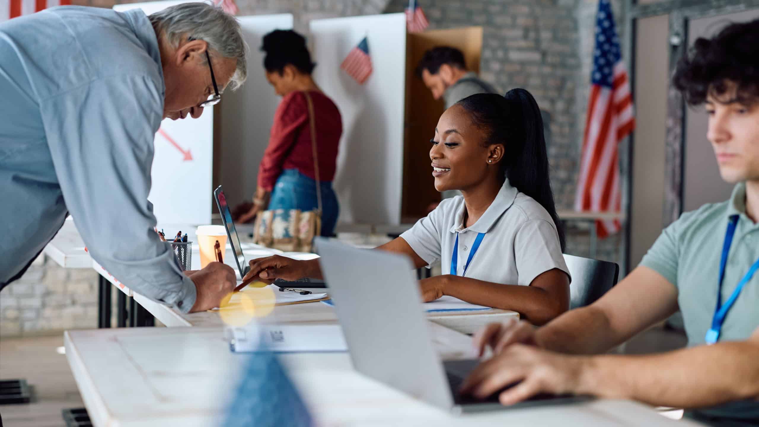 Senior US citizen signing up for voting with help of African American female volunteer at polling place.