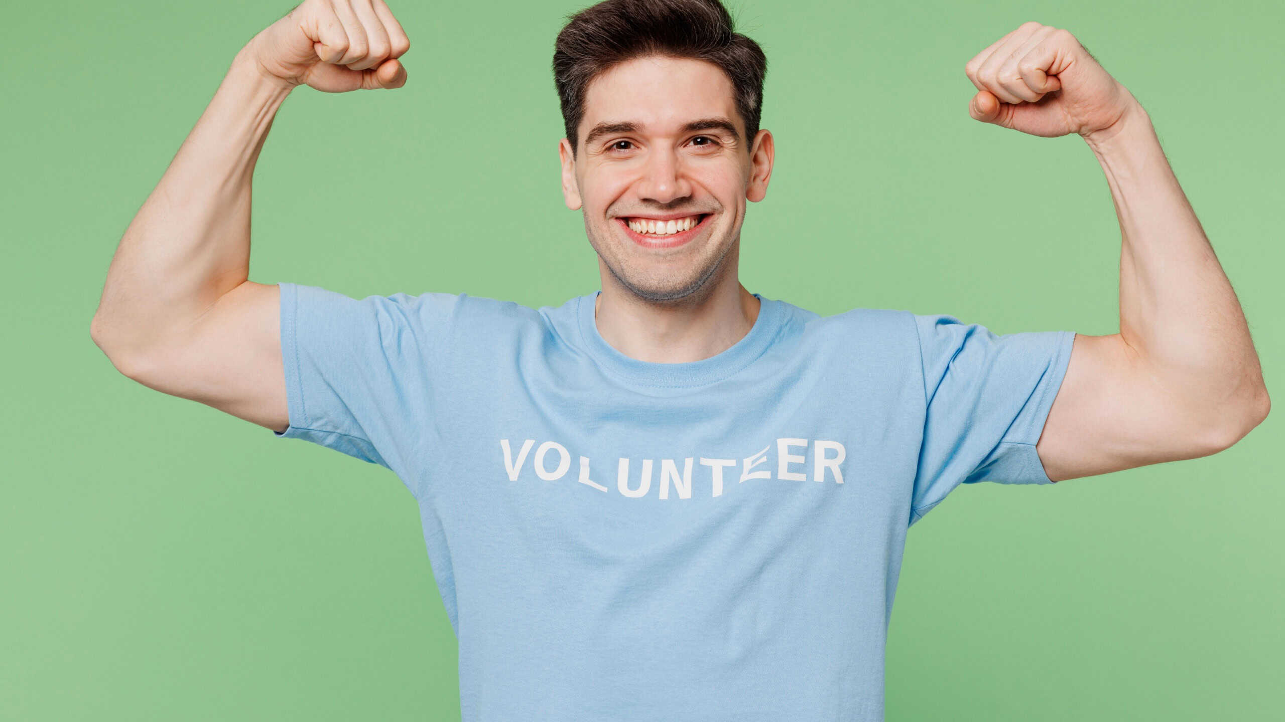 Young man wear blue t-shirt title volunteer show biceps muscles on hand demonstrating power isolated on plain pastel light green background. Voluntary free work assistance help charity grace concept