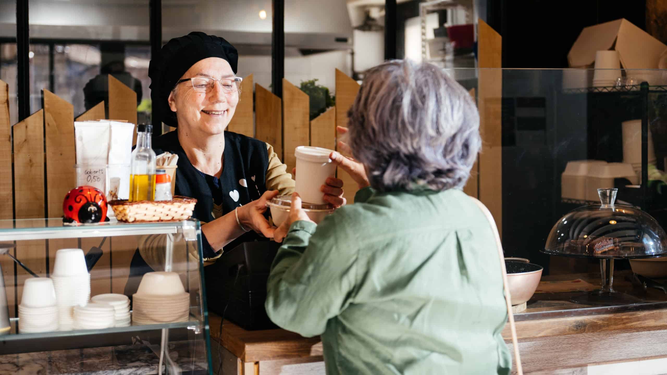 A cheerful woman in a black apron serves a takeaway meal to a customer at a food shop counter, both exchanging smiles.