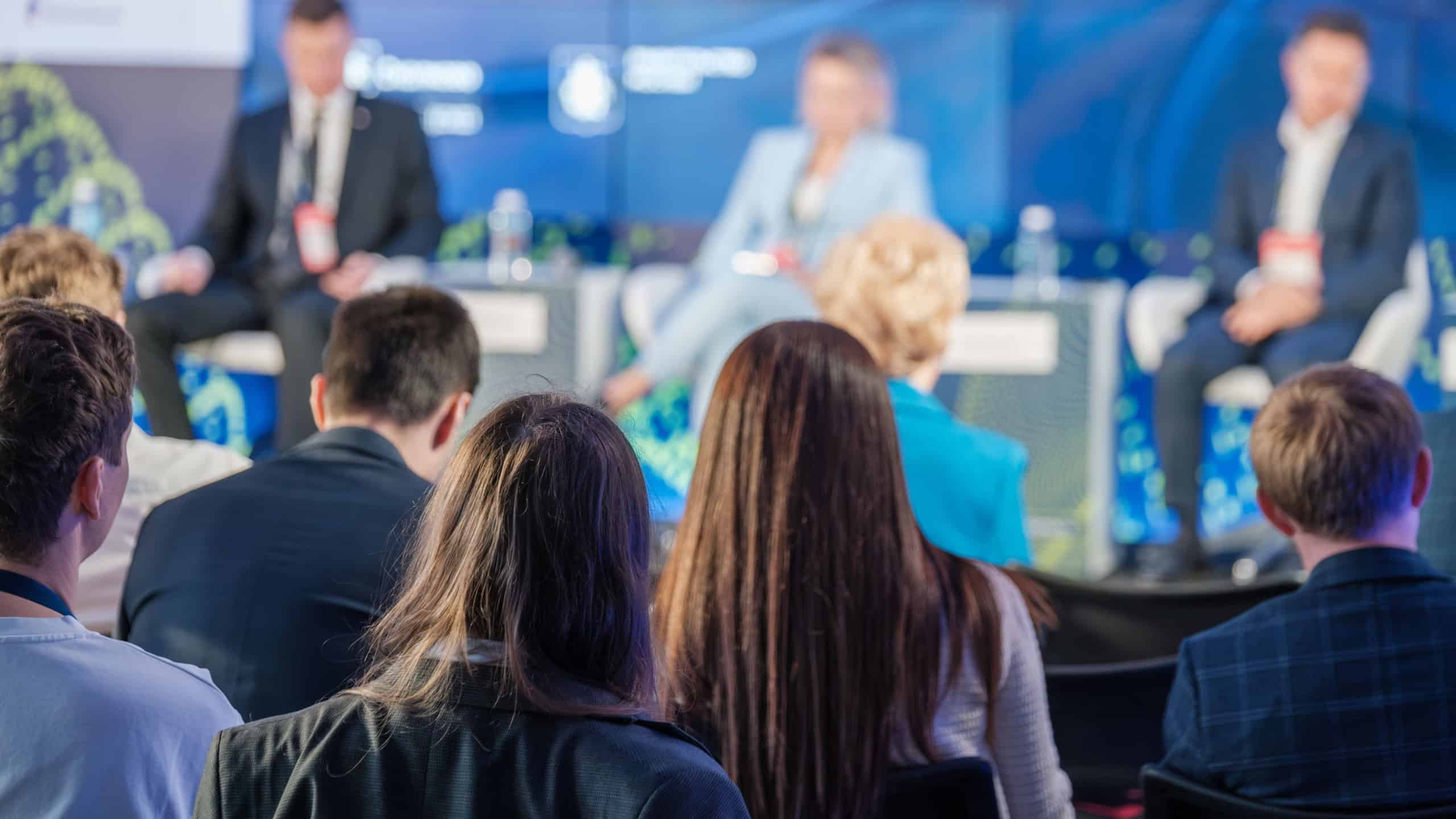 A diverse audience attentively listens to speakers on stage during a business conference. The panelists appear out of focus as they discuss key topics.