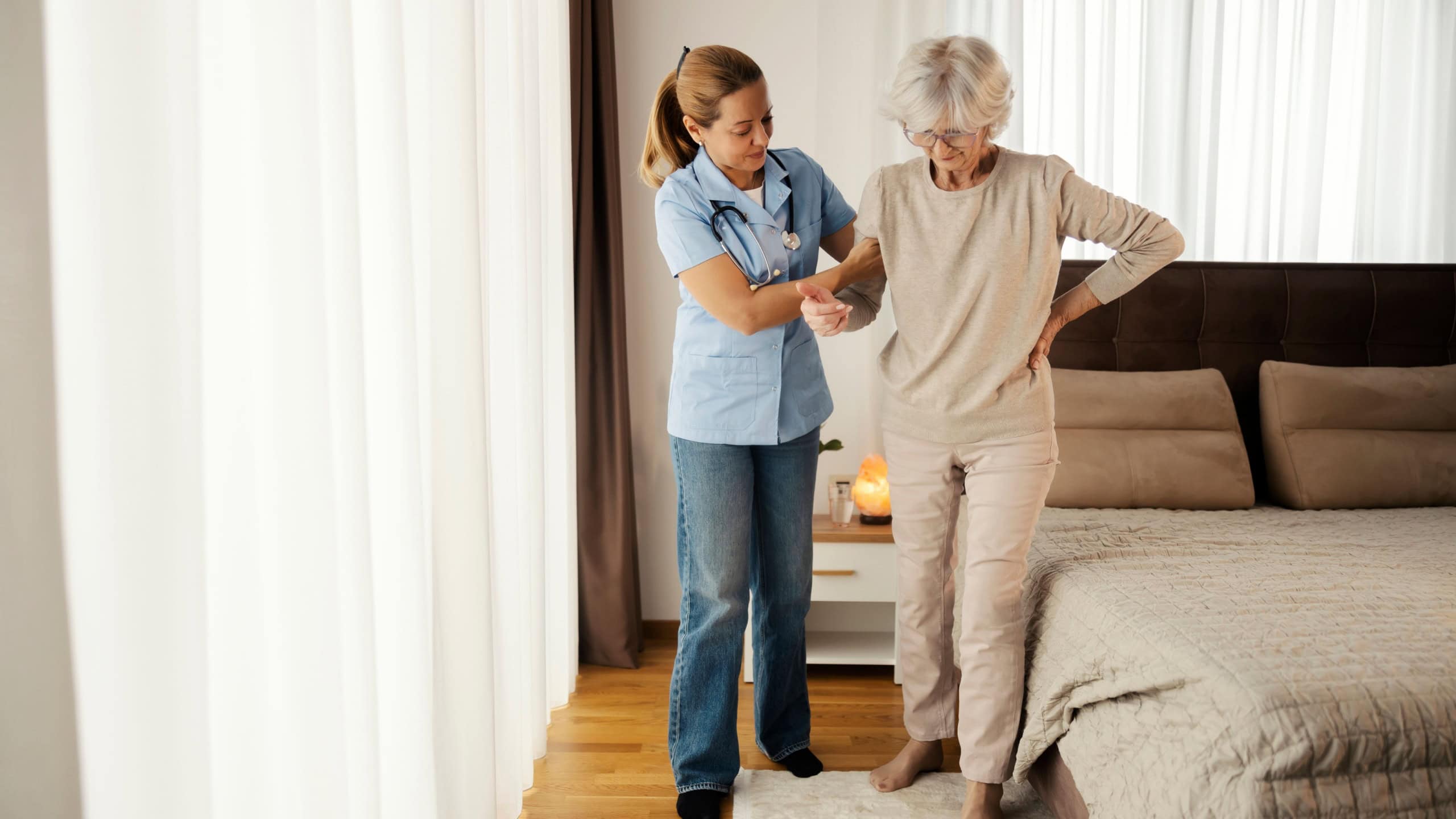 Full length of nurse holding sick old woman under armpit and helping her with walking around bedroom at nursing home.