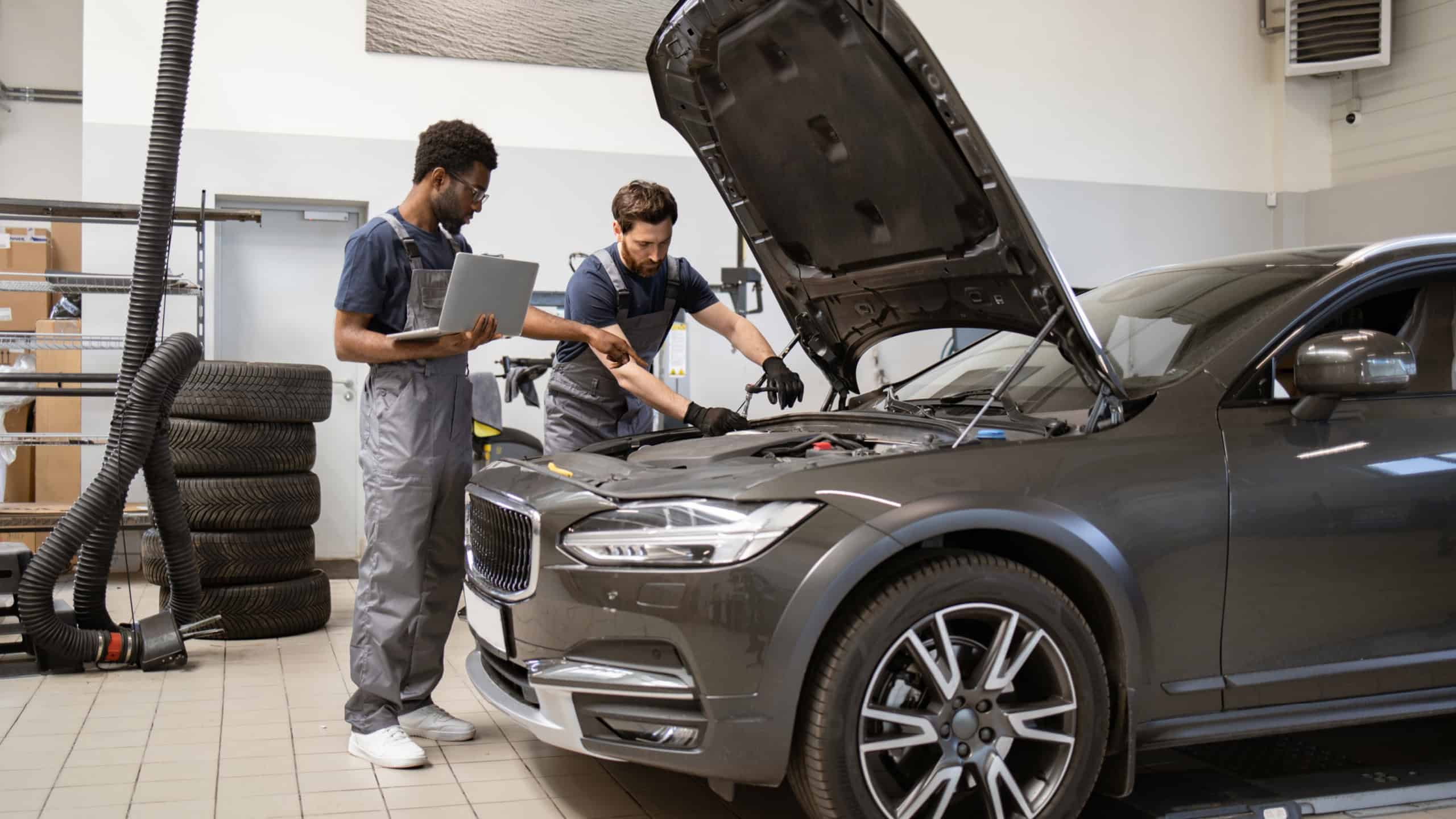 Two auto mechanics inspecting car engine in workshop environment. Professionals diagnosing vehicle with laptop and tools. Automotive maintenance, repair, and teamwork concept