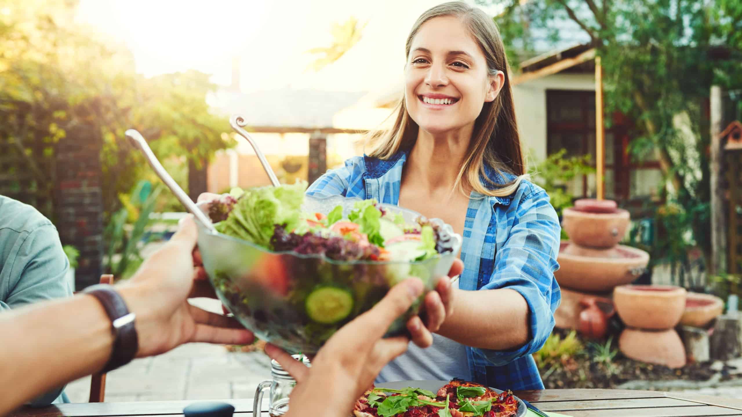 Social, happy woman and salad in backyard for lunch, sharing or healthy meal in summer. Female person, food and friendship on estate for birthday party, reunion and smile at outdoor table with people
