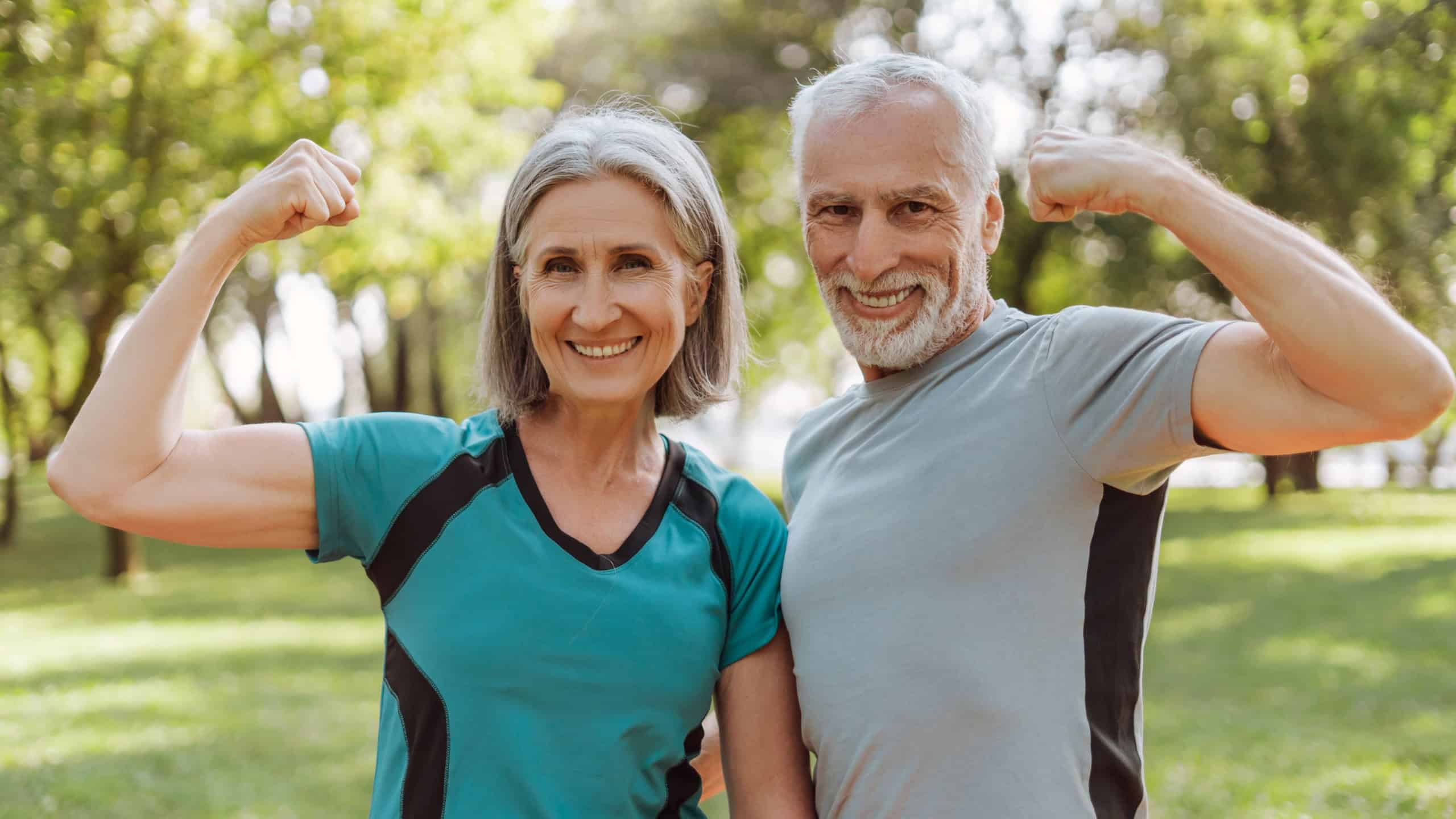 Smiling, attractive senior athletic couple flexing muscles in park, looking at camera outdoors, active lifestyle concept