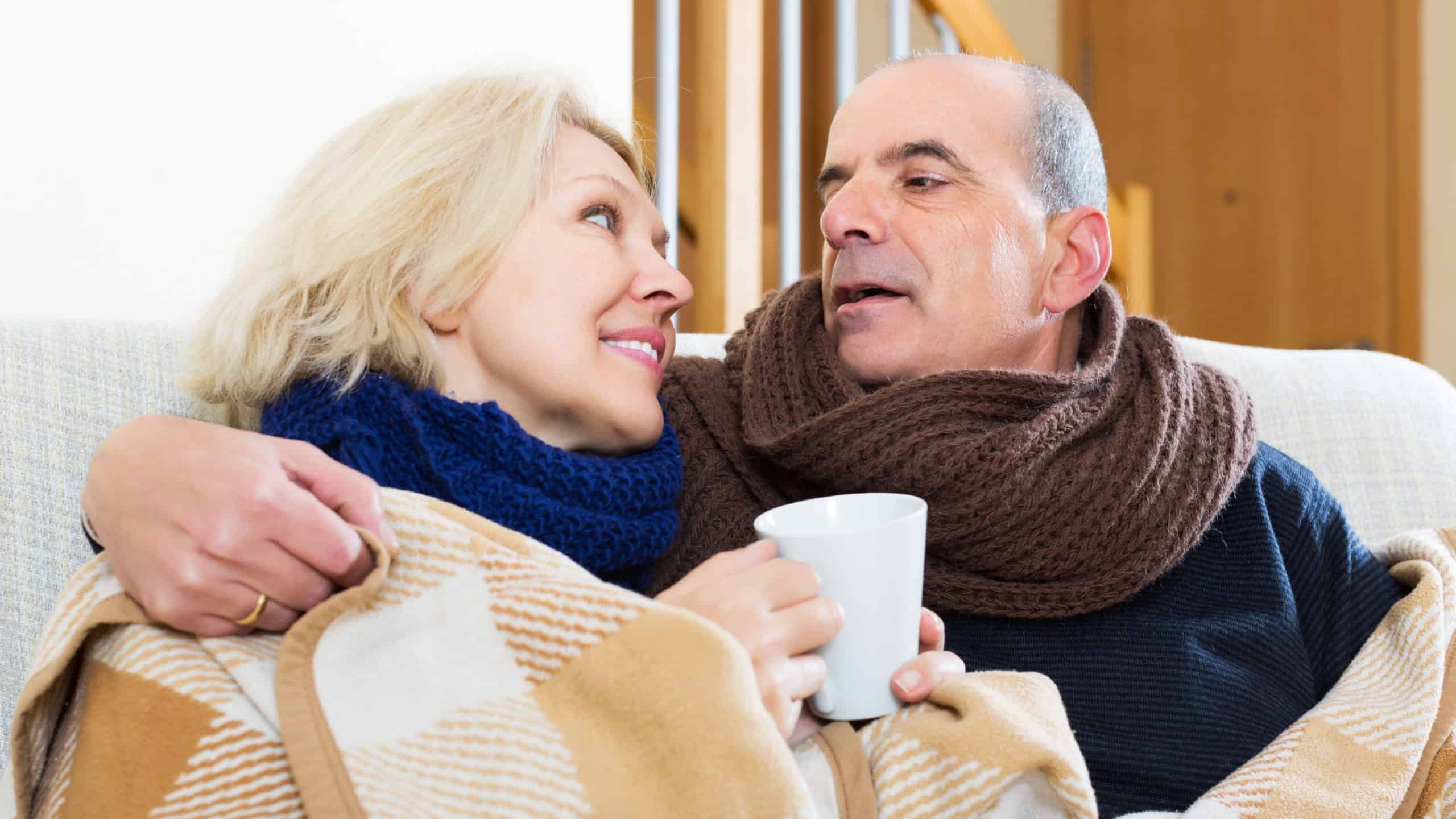 Elderly smiling spouses under blanket drinking tea on couch