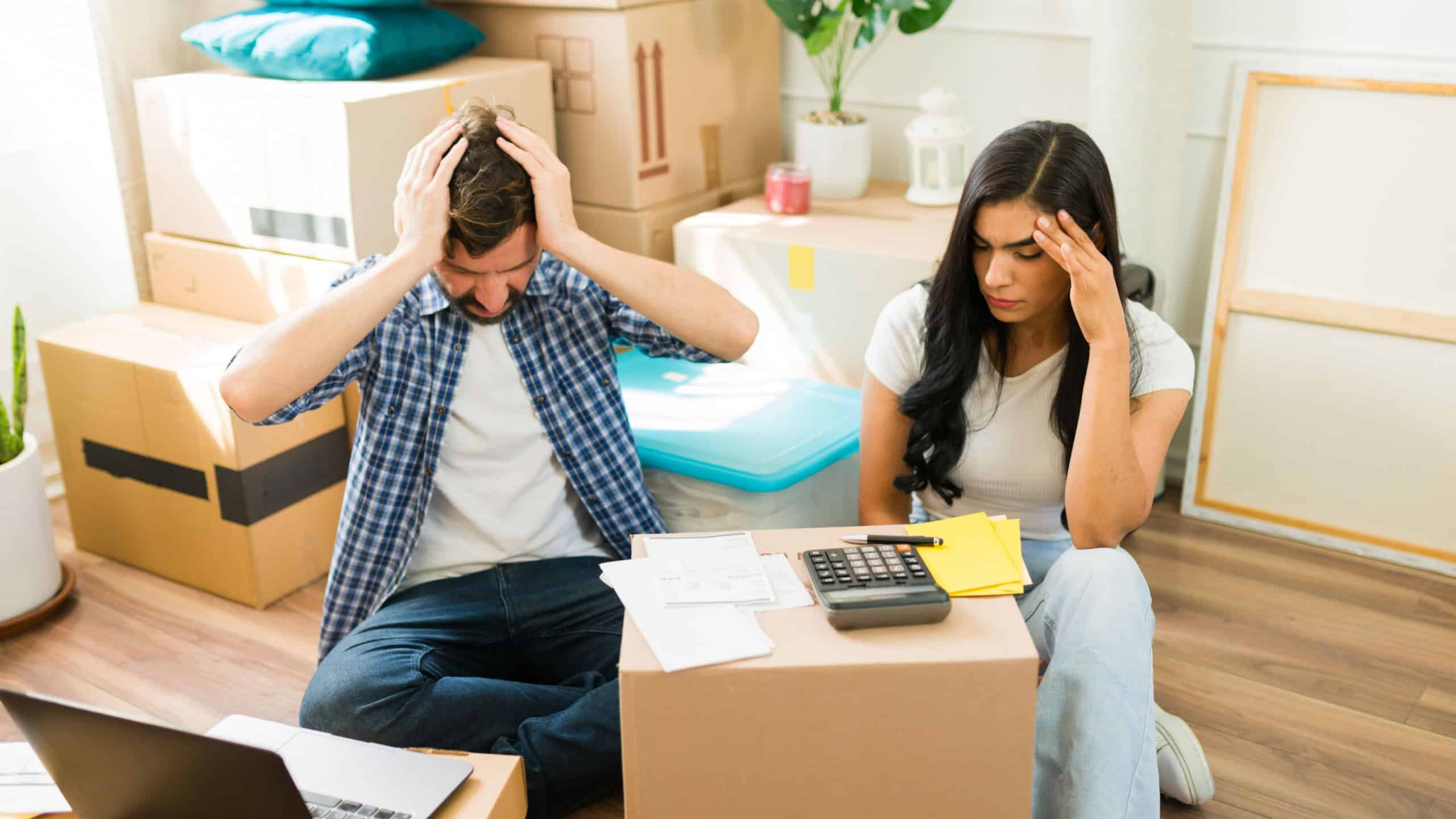 Young couple is sitting on the floor surrounded by moving boxes and feeling stressed while planning their budget in their new home