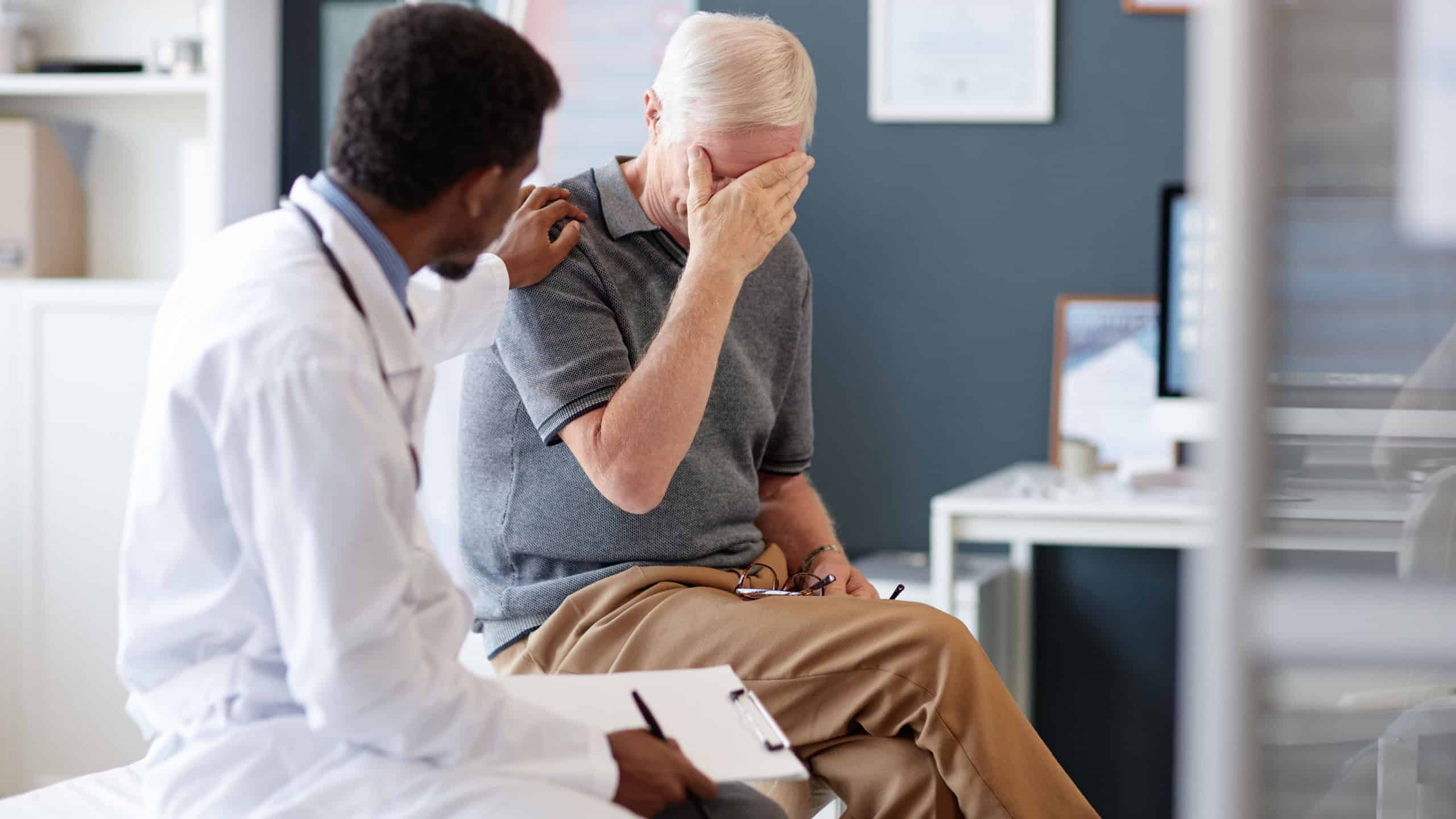 Candid scene with Caucasian senior man crying during consultation in clinic and covering face while doctor comforting patient