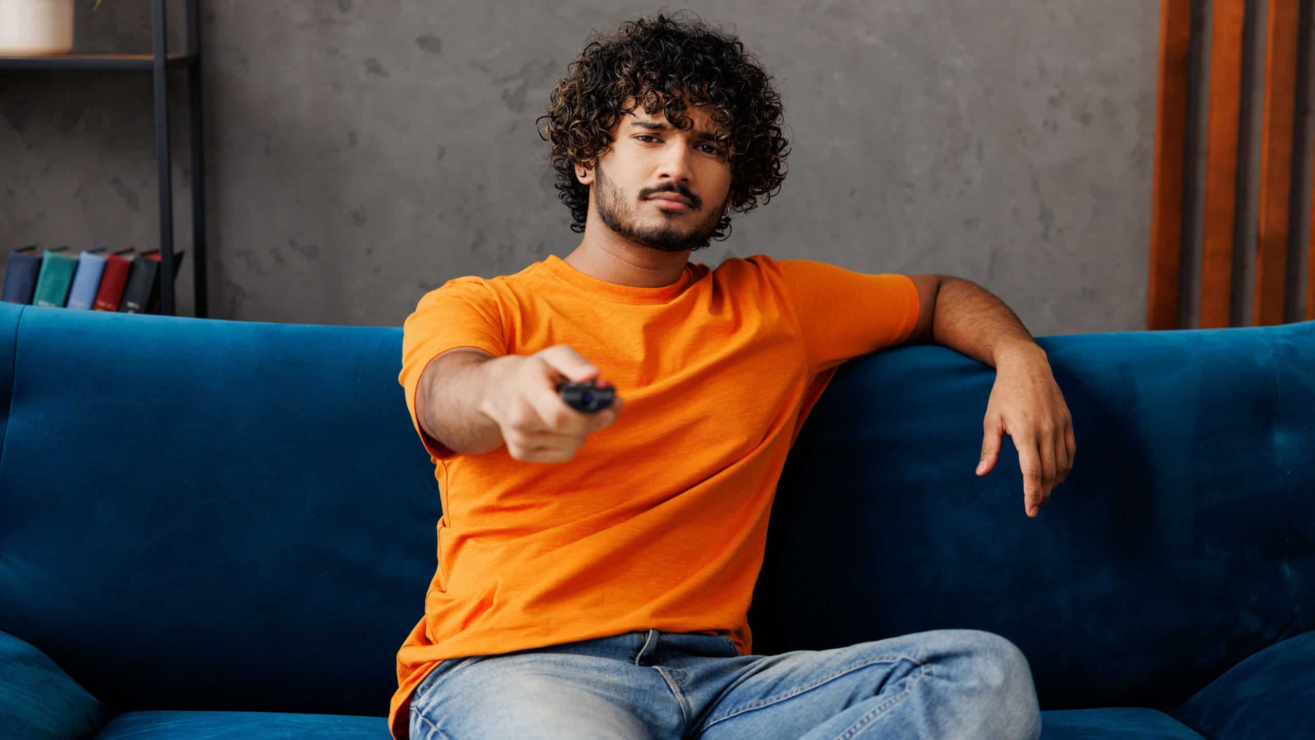 Young frustrated bored Indian man wear orange casual clothes sits on blue sofa couch stay at home switch channel watch tv hotel flat rest relax spend free time in living room indoor. Lounge concept