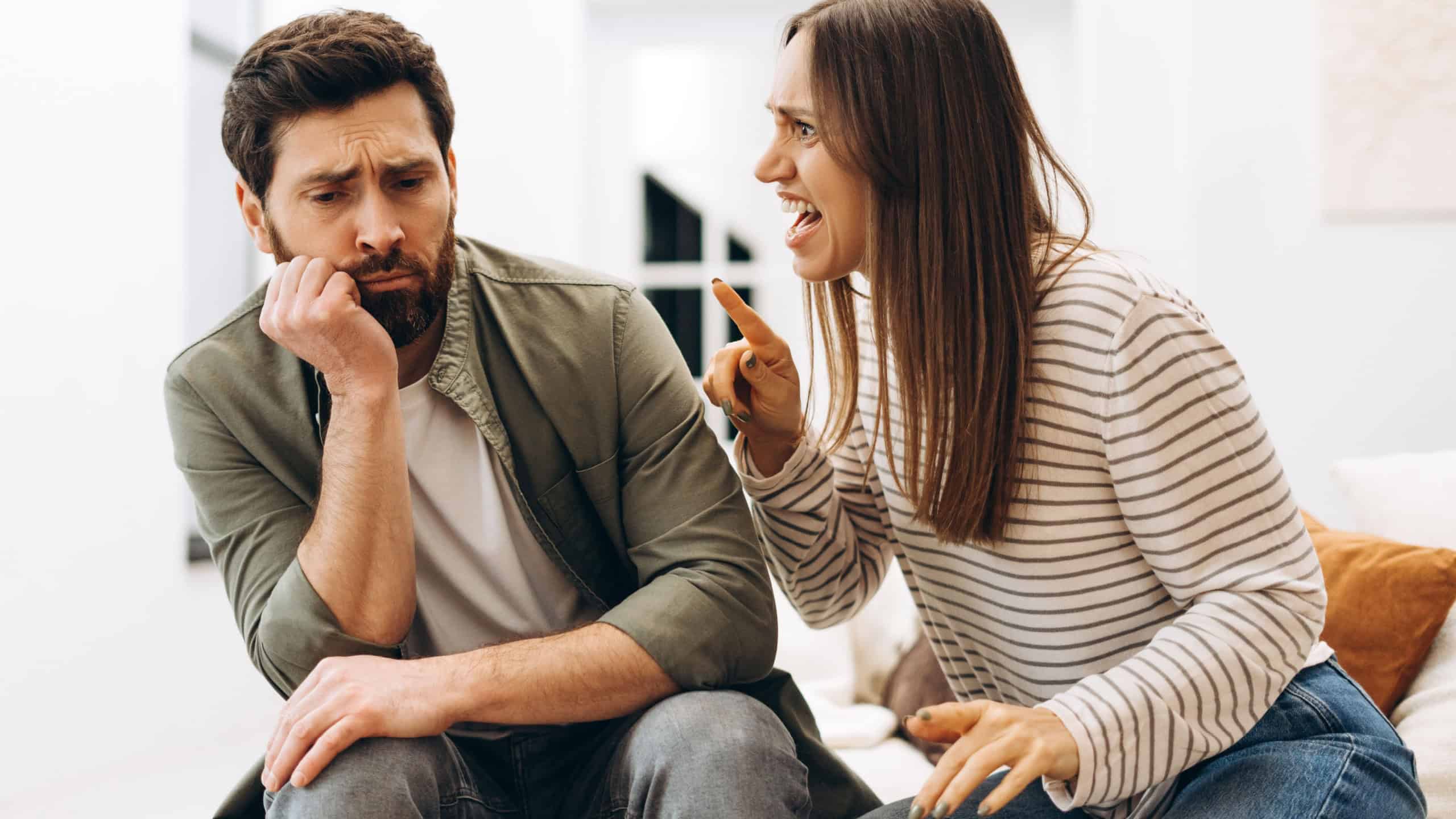 Young woman arguing and screaming at her sad husband sitting on the sofa at home, he looks resigned and is touching his beard with his hand
