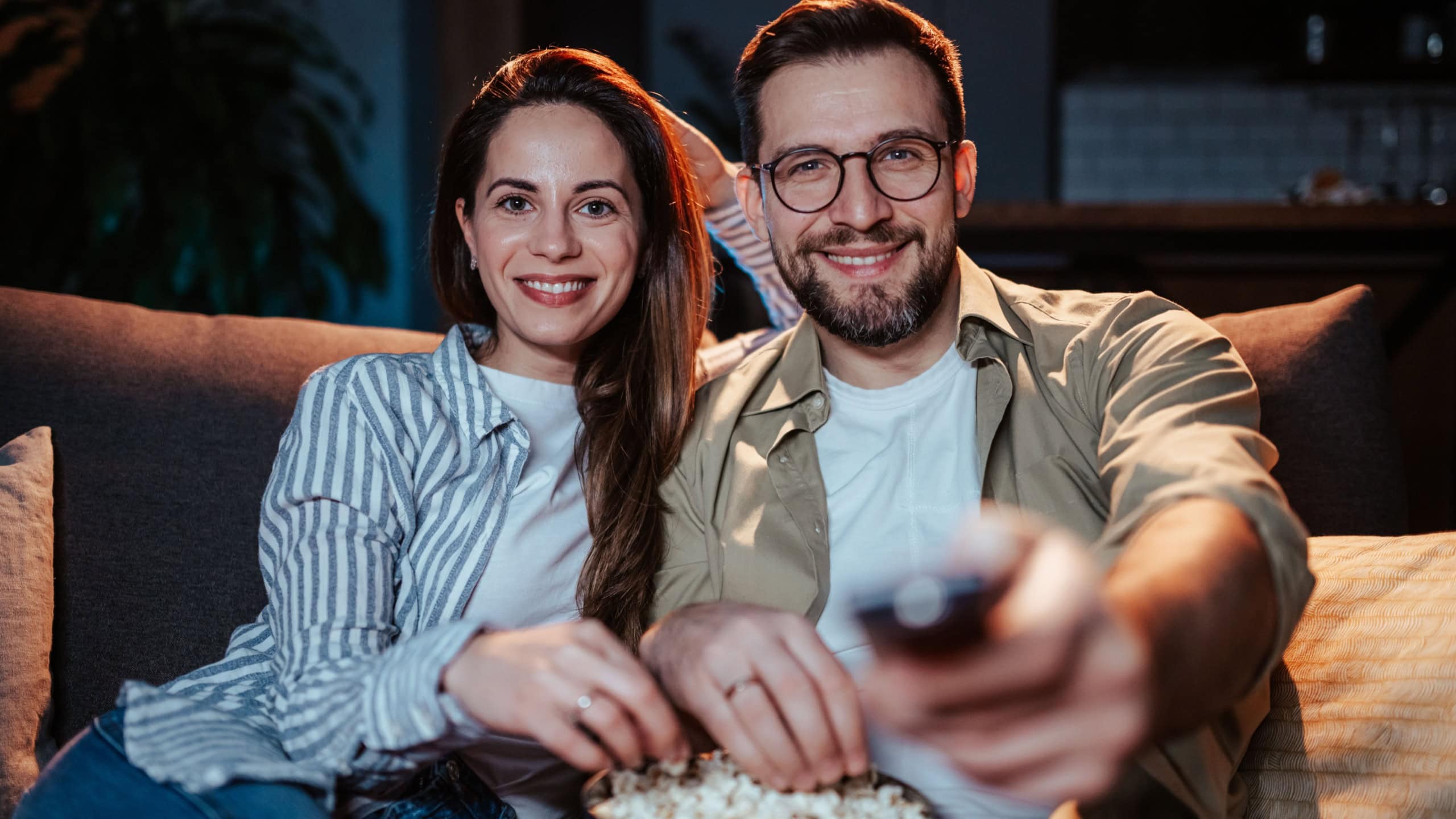 Smiling couple relaxing on a cozy sofa, sharing a bowl of popcorn while watching tv at home. The warm ambiance suggests a comfortable evening spent together in leisure