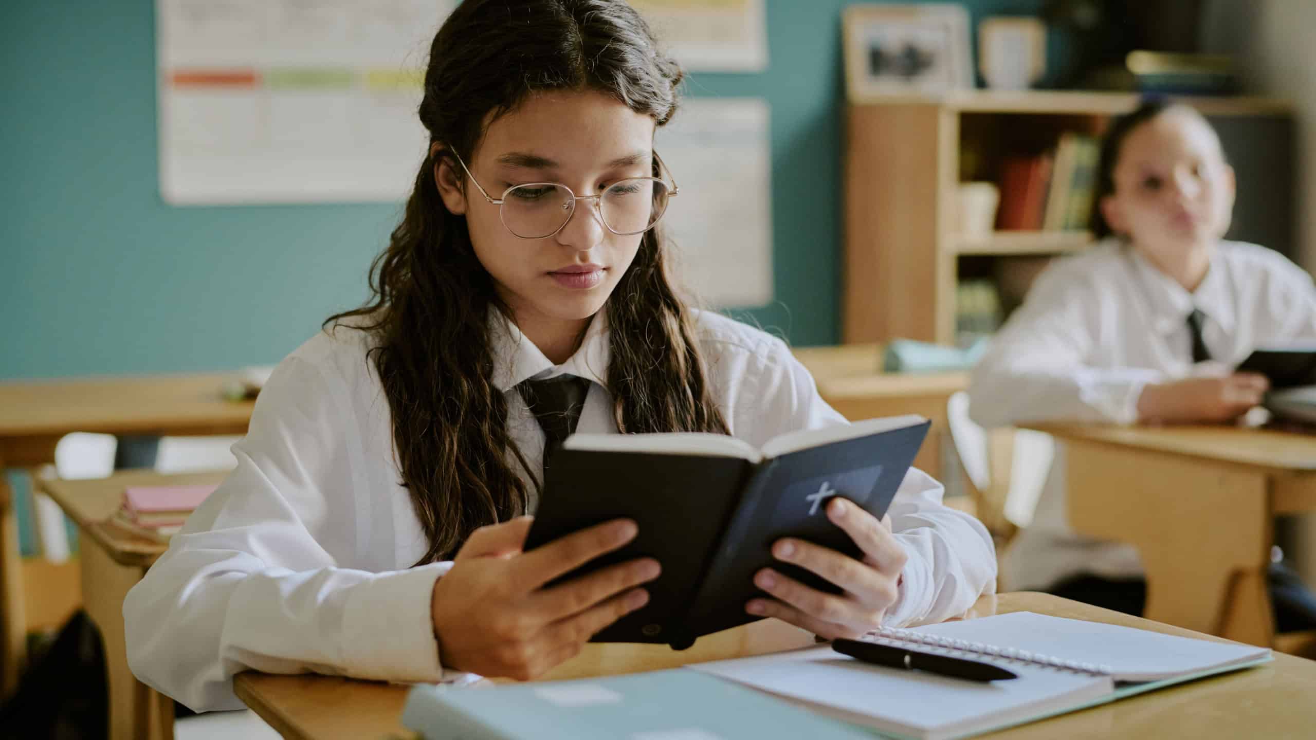 Student with glasses intensely reading Bible in classroom. Setting including open notebooks on desks and another student in background