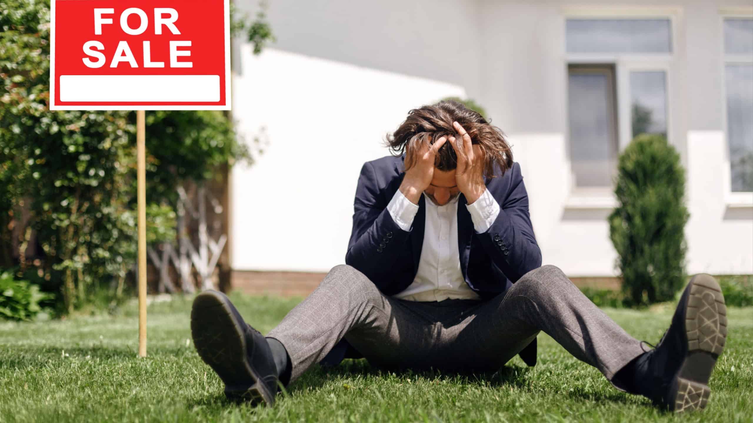 Depressed real estate agent sitting on grass in front of house for sale, outdoors