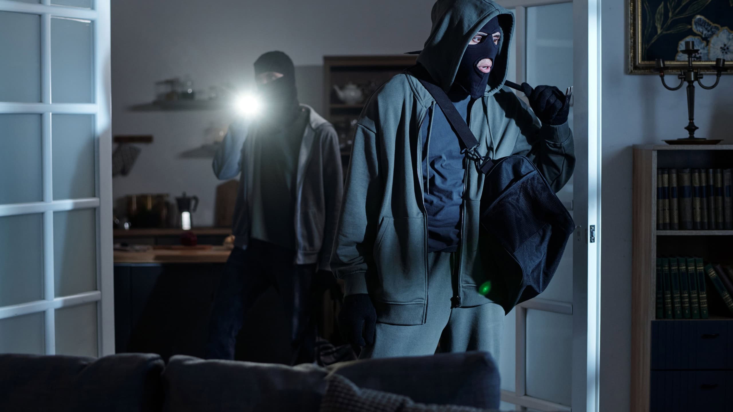 Two masked men entering dark room, one carrying large bag and other shining flashlight, both wearing hooded clothing and gloves, appearing to commit burglary