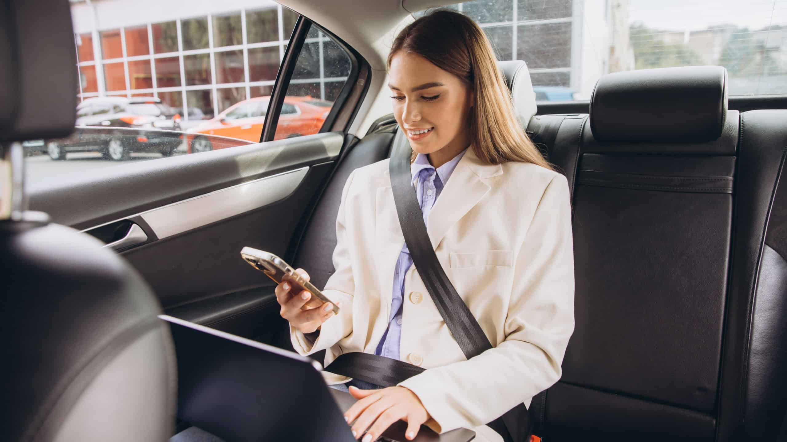 A young woman in professional attire works on her laptop and uses a smartphone while sitting in the backseat of a car, multitasking efficiently during travel.