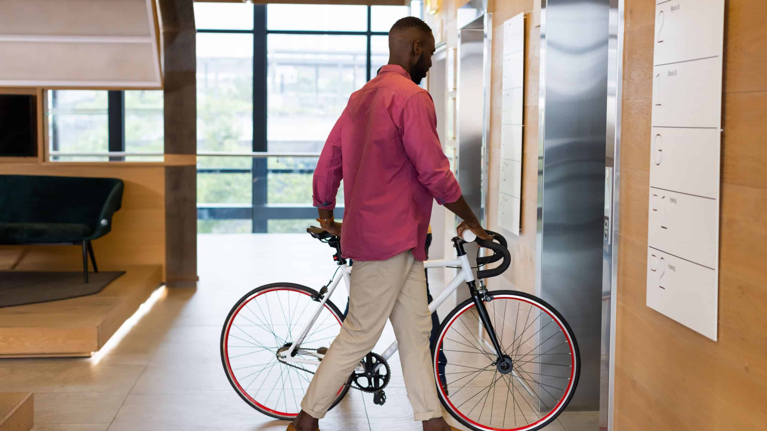 African American male in magenta shirt pushing road bicycle through lobby near elevator doors. Contemporary, urban, lifestyle, architecture, transportation, leisure, vibrant