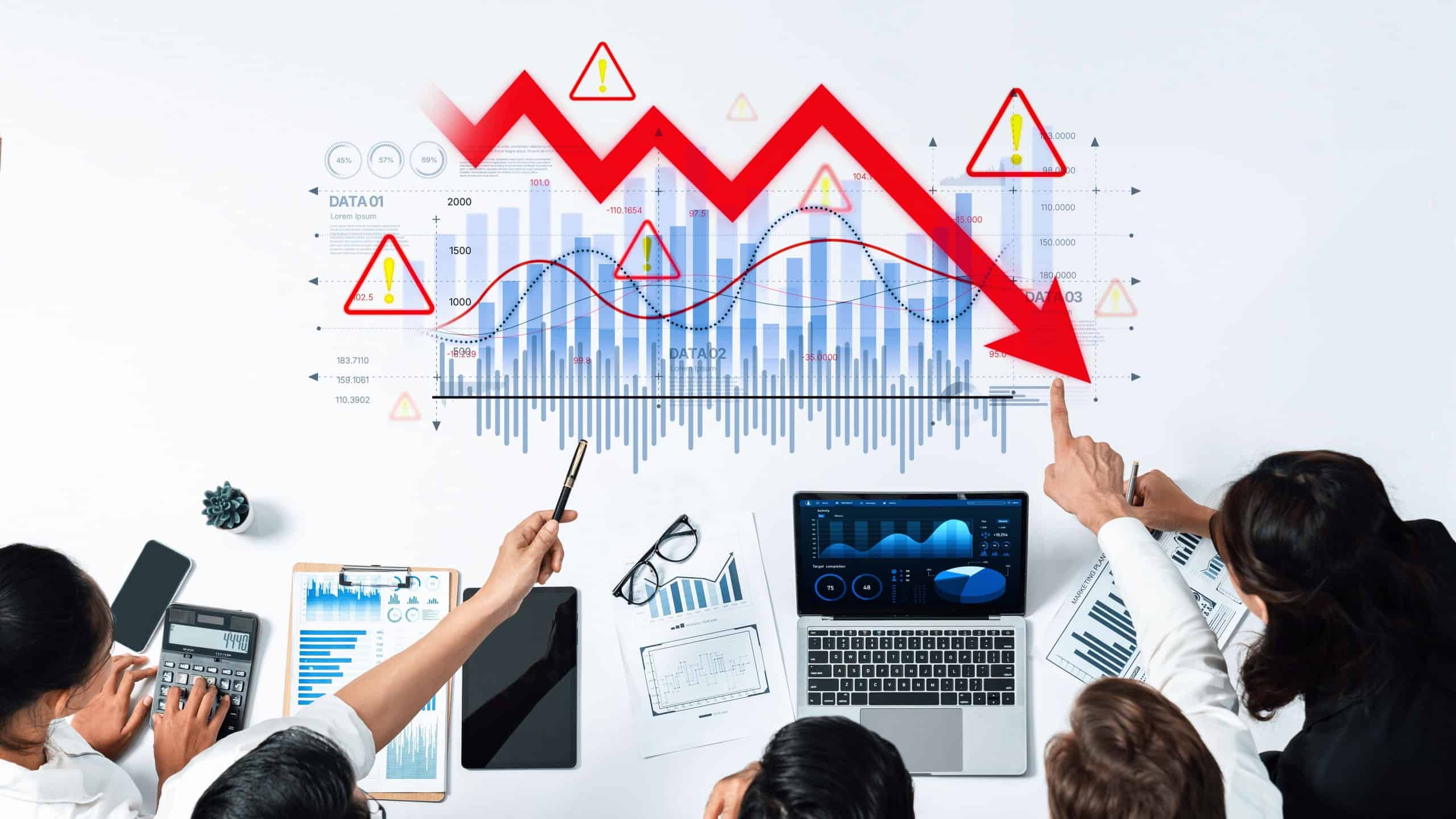 Professionals collaboratively analyzing financial data with declining graphs and warning signals, highlighting market trends, challenges, and strategic planning efforts.