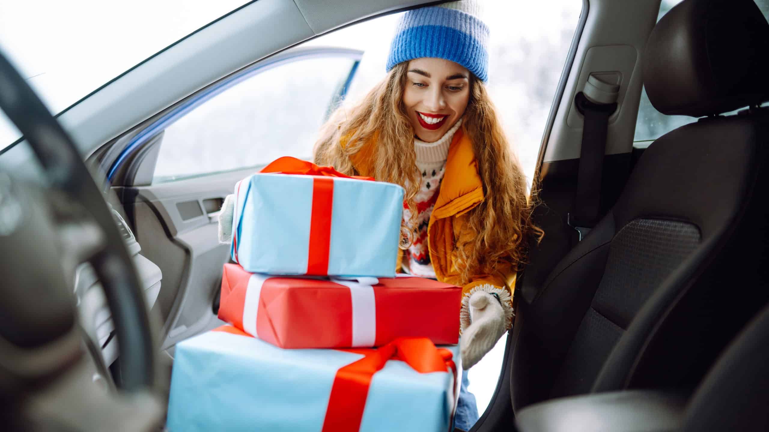 Happy woman in bright jacket packs wrapped Christmas gifts into her car on a sunny winter day. Cute woman enjoys snowy day and prepares for New Year's holidays. Holiday and weekend concept.