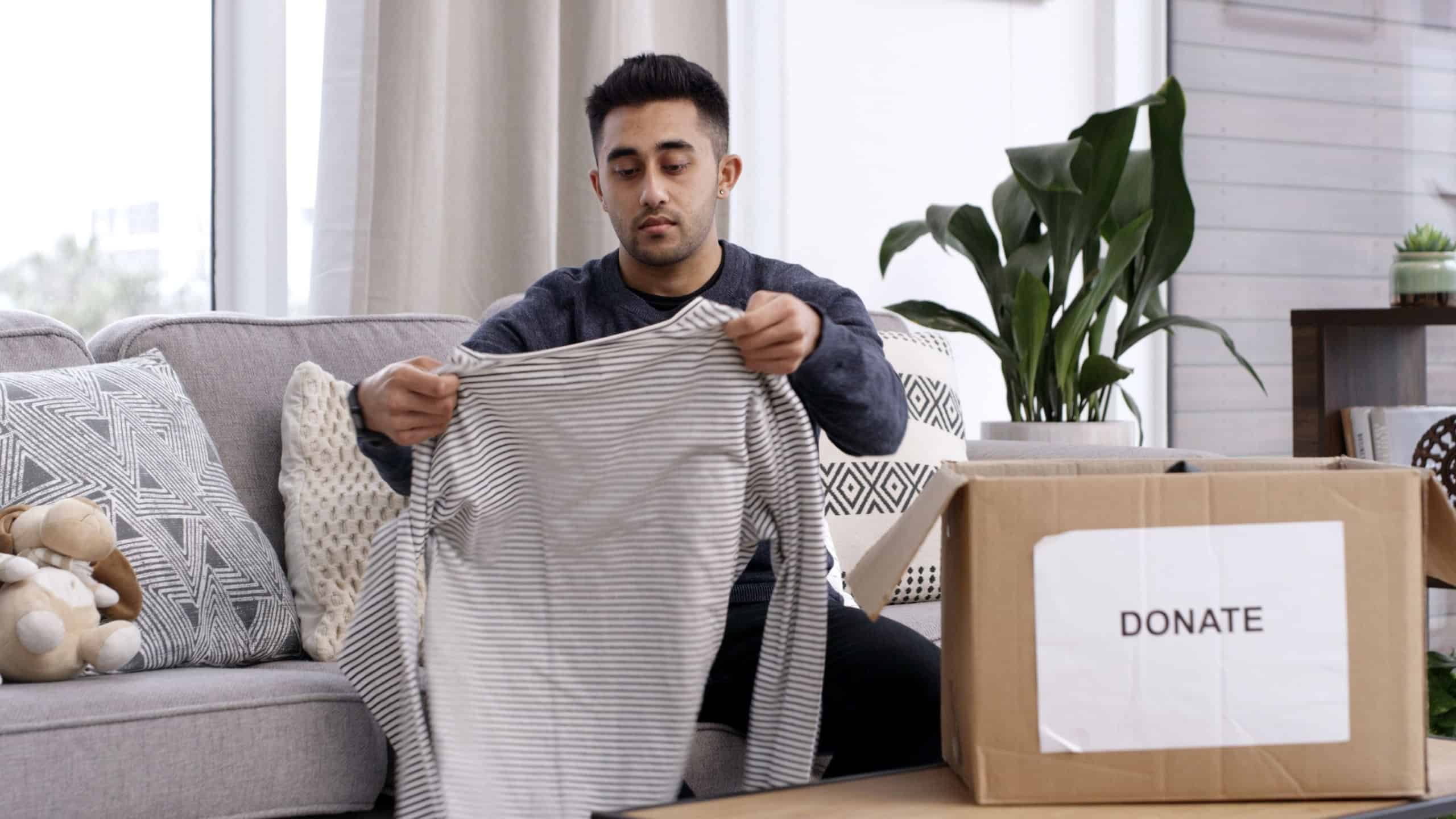 Man, folding clothes and donation box in house for charity initiative, nonprofit and declutter closet. Person, package and apparel giveaway in home for NGO contribution, support and spring cleaning.