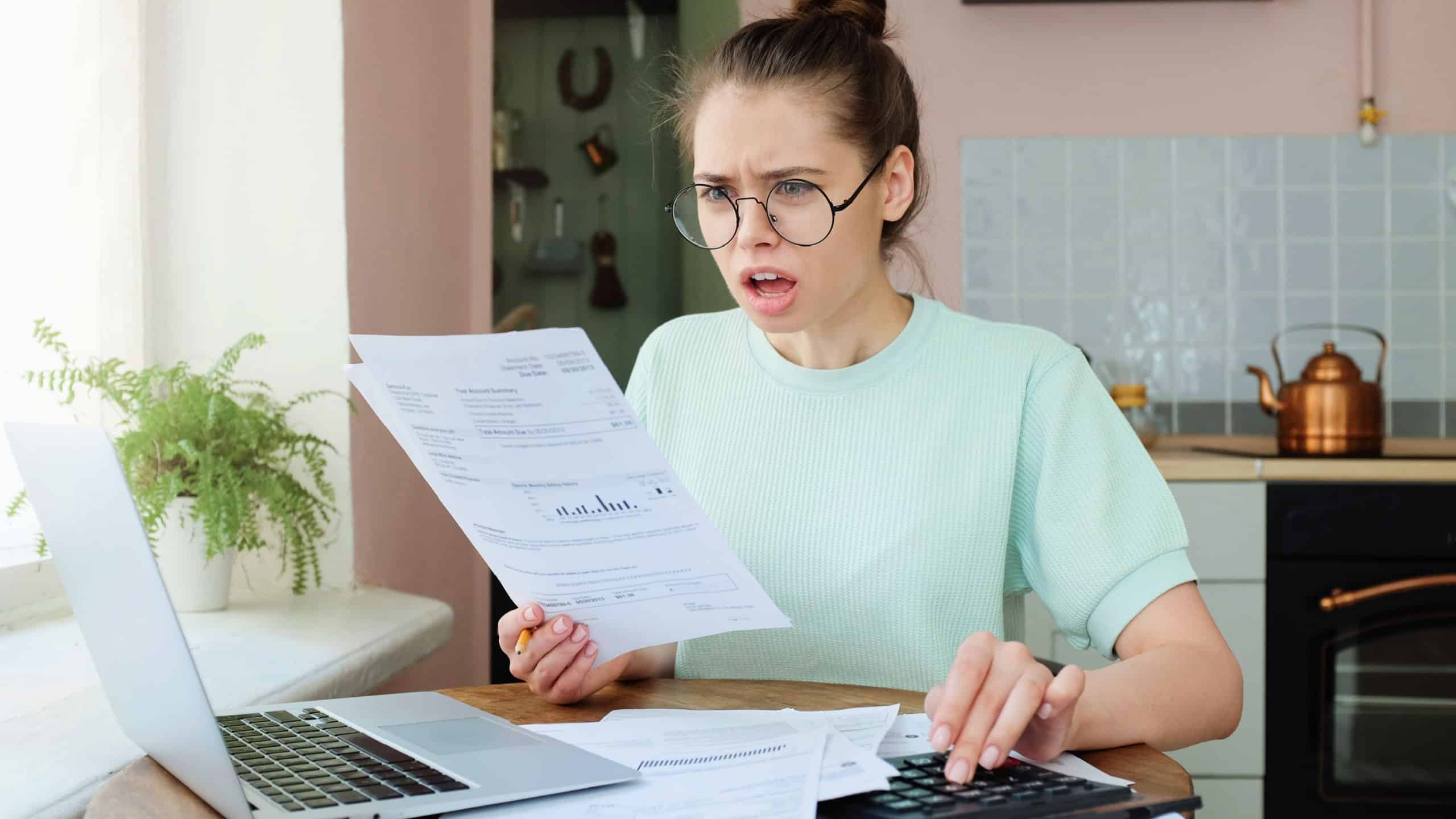 Indoor closeup of young emotional European lady dealing with sheets of paper and bills at home having opened mouth as if she can not believe these big numbers to pay are true, feeling despair