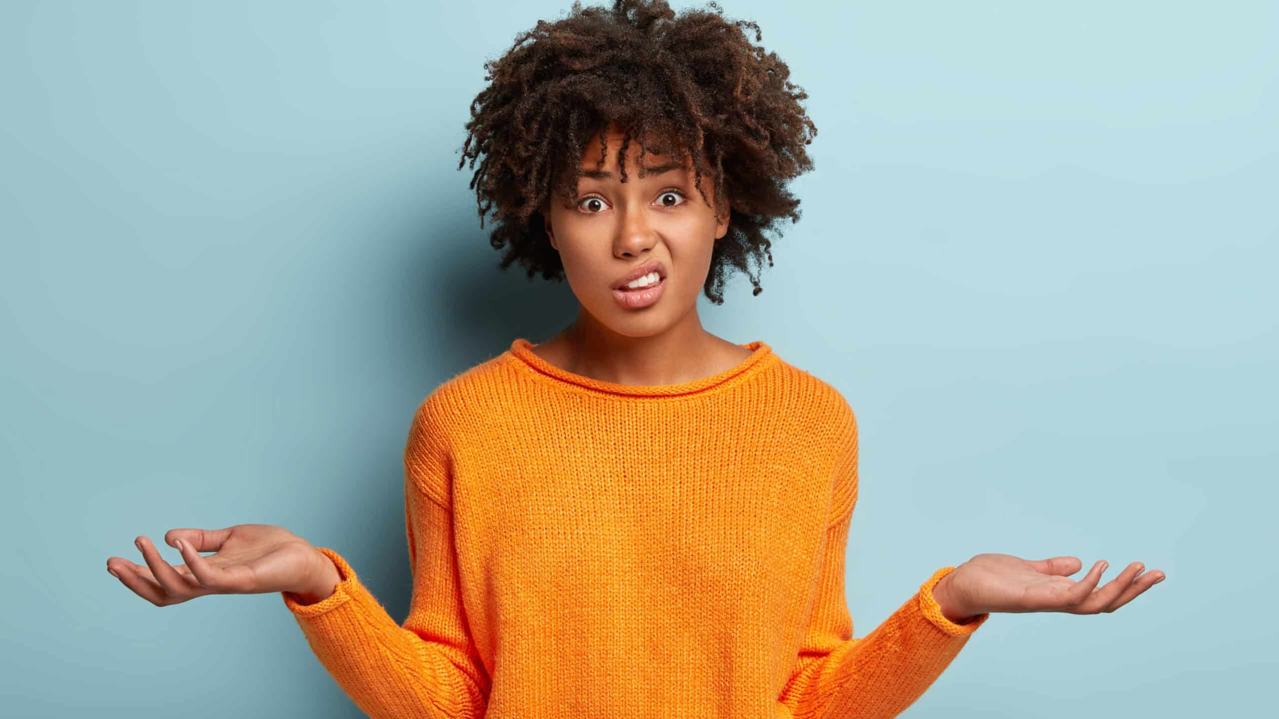 Horizontal shot of attractive woman with puzzled expression, spreads hands with confused look, frowns face, wears orange clothes, feels hesitant, indignant isolated over blue background. Doubt concept