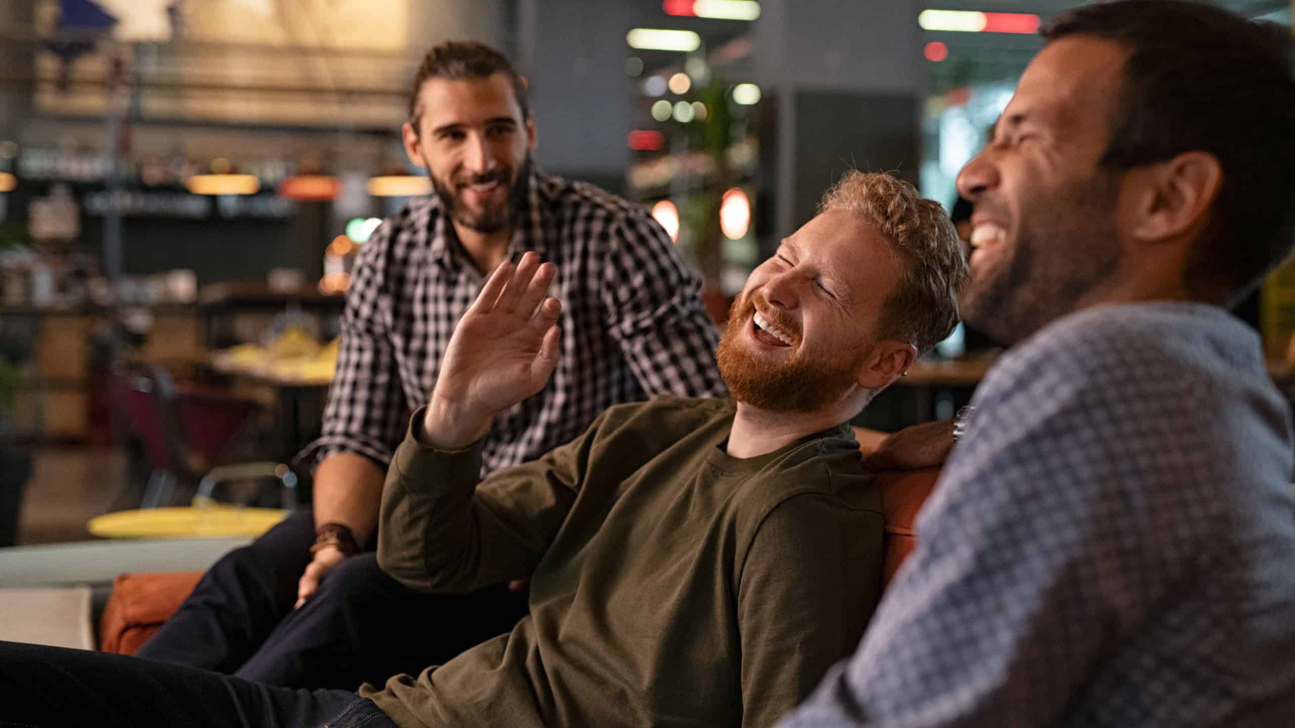 Group of three best friends laughing and enjoying the evening at pub. Happy young men enjoying late night staying together at bar. Cheerful guys sitting on couch and having fun while relaxing.
