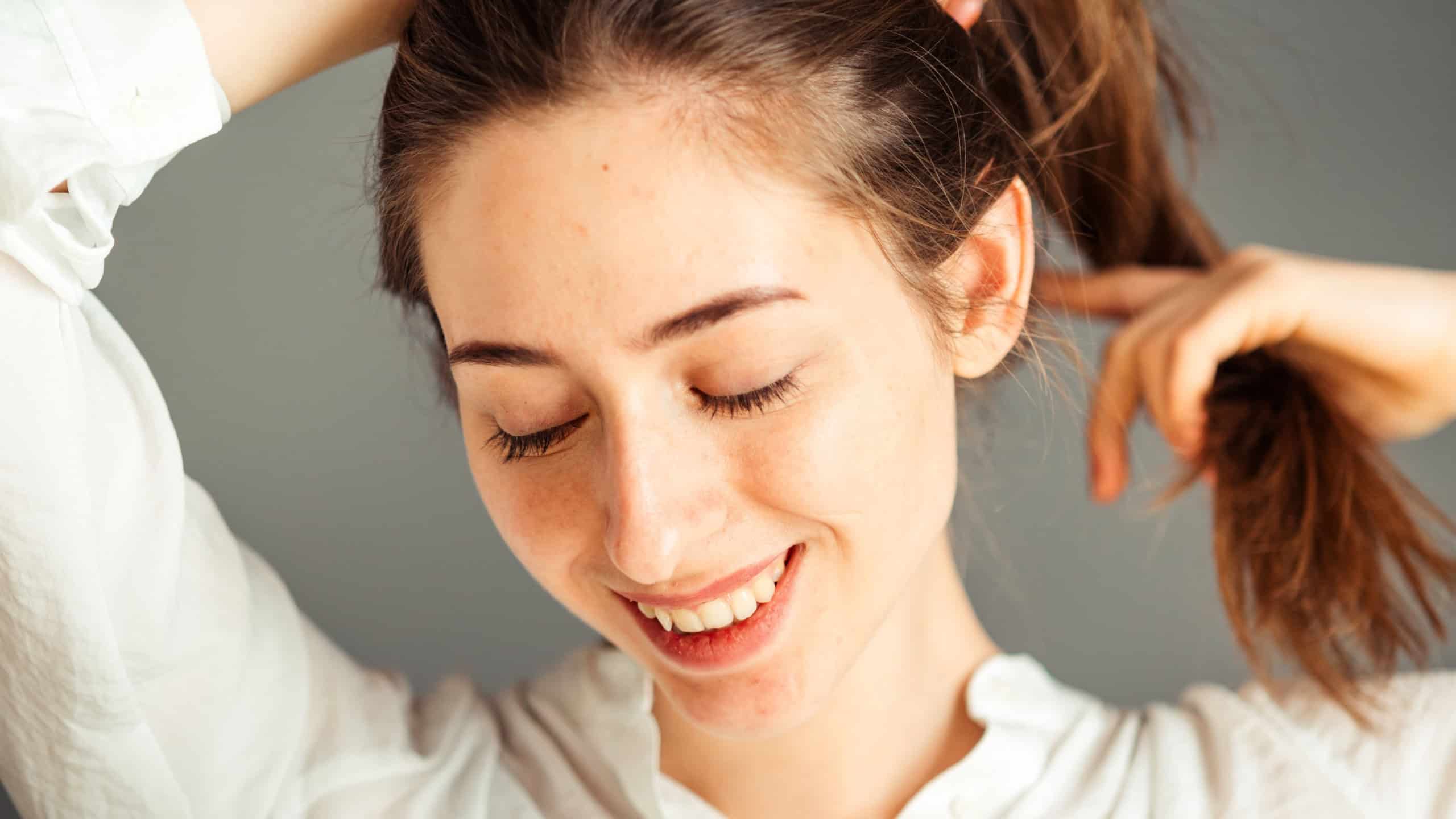 A smiling girl with a satisfied face holds her hair with her hands on a gray background, close-up. Nude makeup, without retouching.