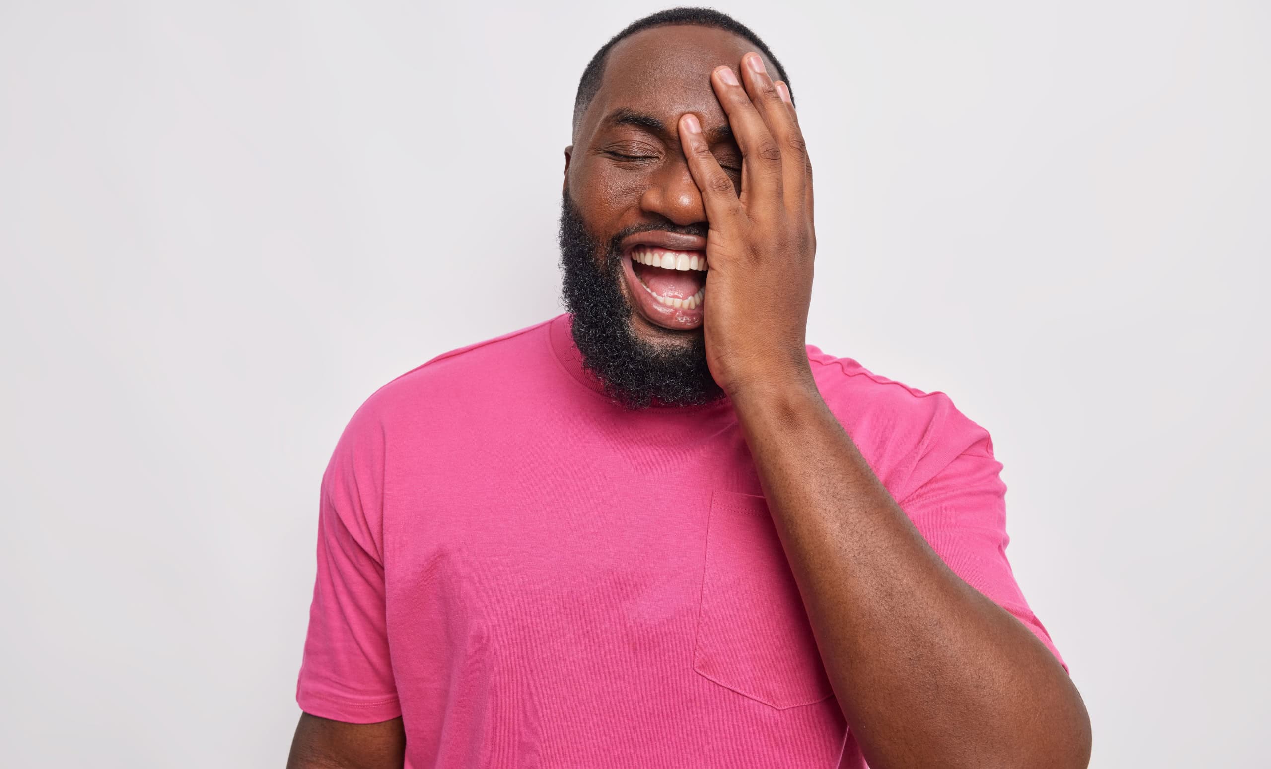 Happy dark skinned man makes face palm keeps eyes closed smiles carefree dressed in casual pink t shirt laughs at funny joke isolated over white background. Human emotions and feelings concept