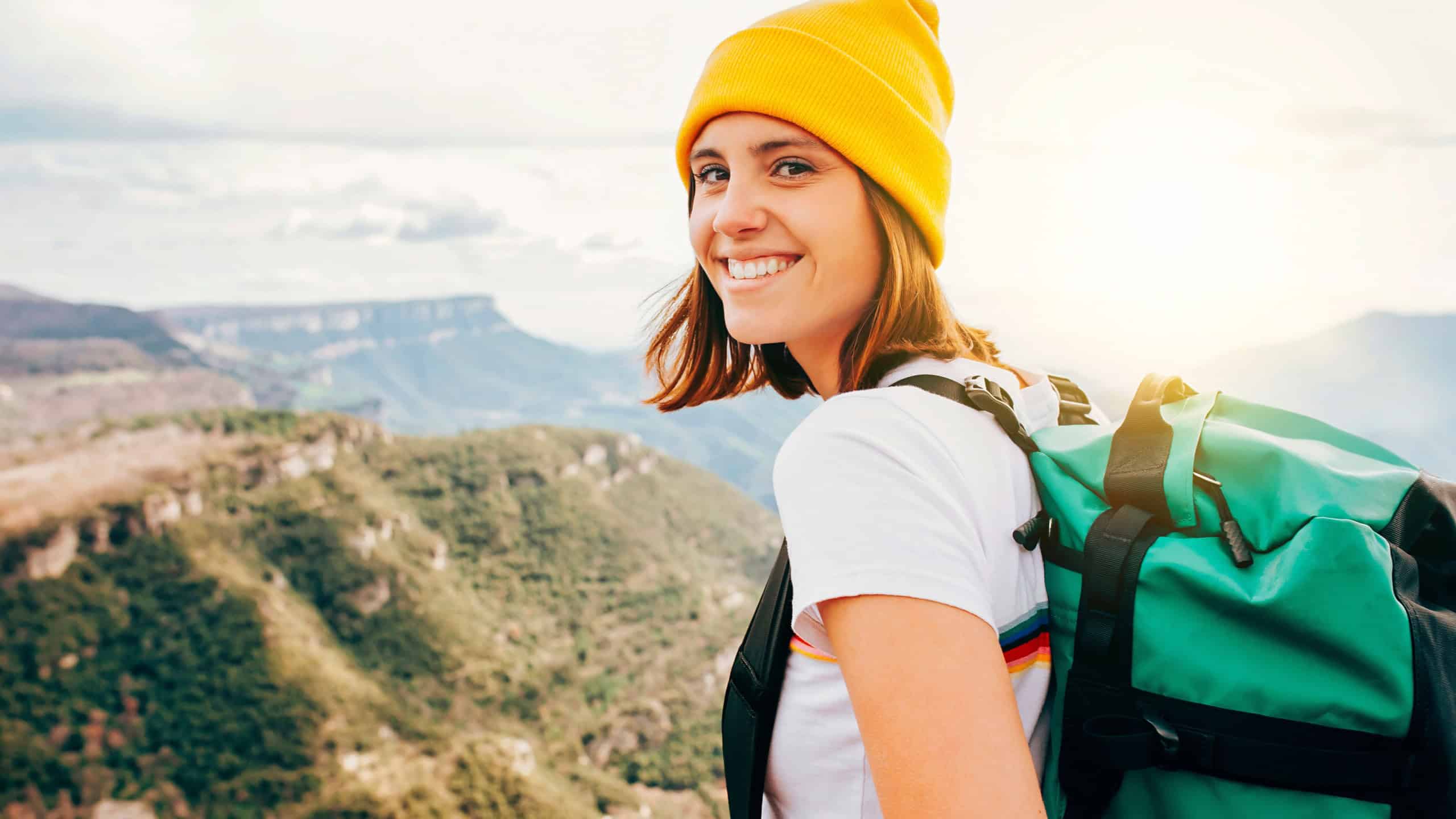 Back view young happy smiling woman backpacker with brunette hair relaxation on top rocky mountains, with backpack, yellow hat. Leisure after walking valley. Adventure, travel, holiday concept.