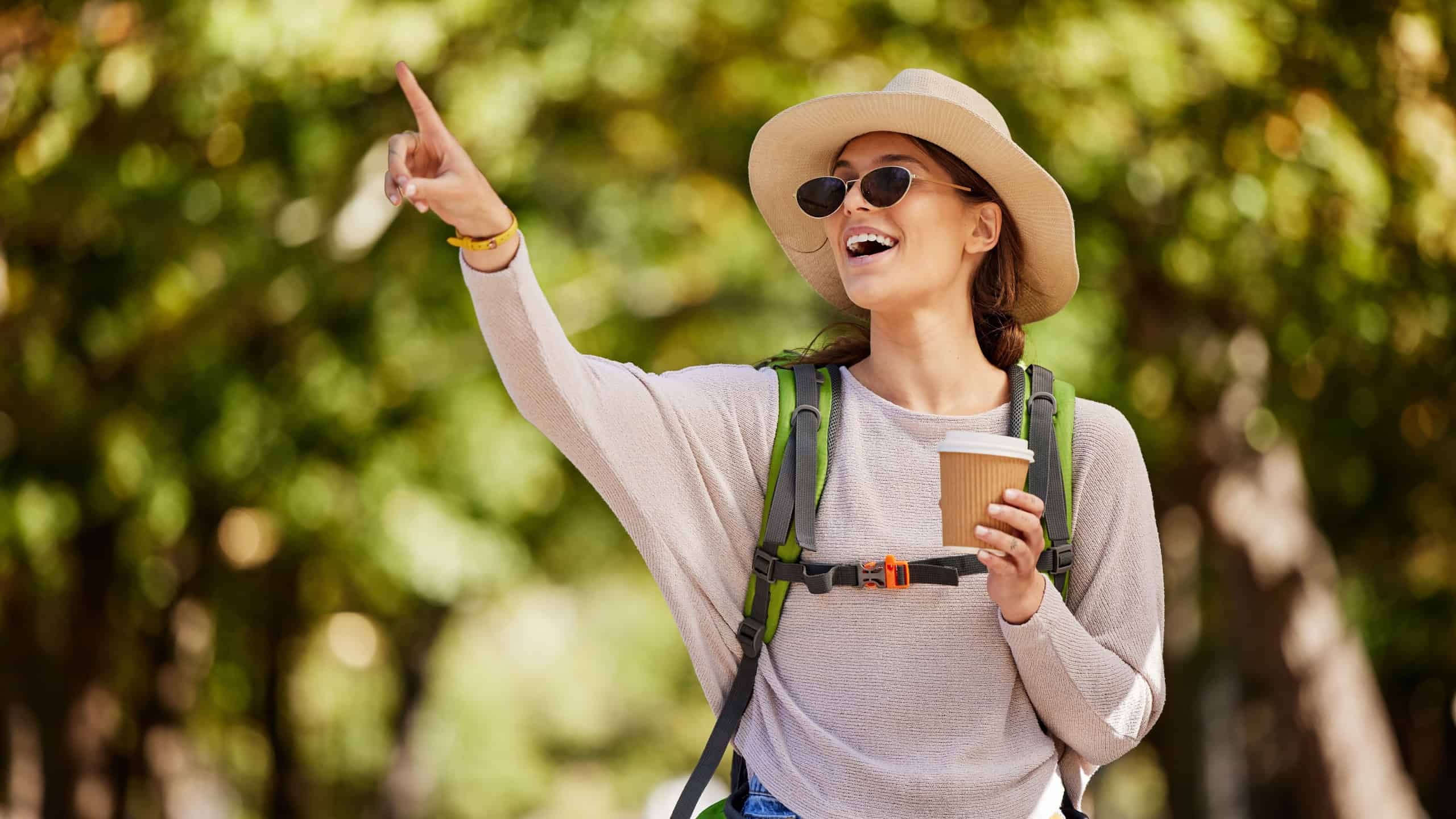 Woman, backpack or pointing in nature travel, sightseeing park or bird watching adventure in trees forest or Canada woods. Smile, happy tourist and showing hand gesture in hiking location with coffee