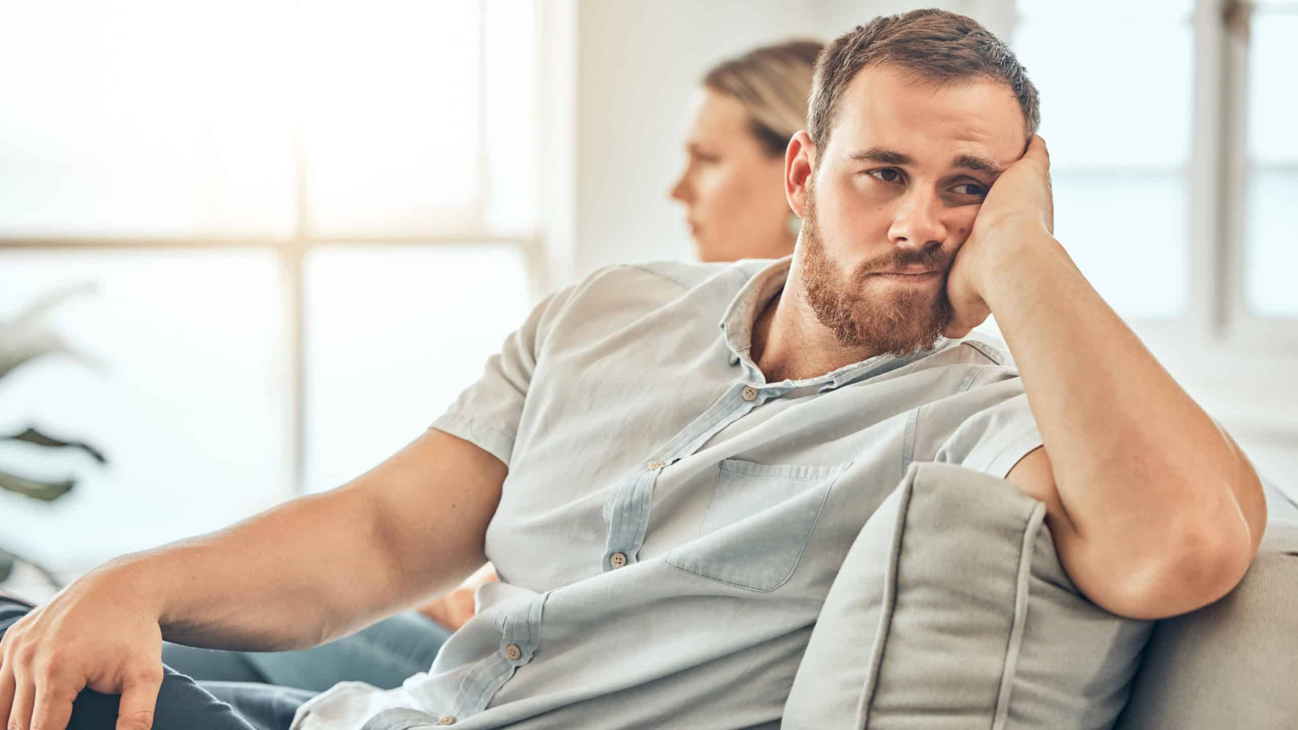 Young caucasian man with a beard looking unhappy and annoyed while sitting on the couch during an argument with his wife at home. Bored man sitting and thinking on the couch.