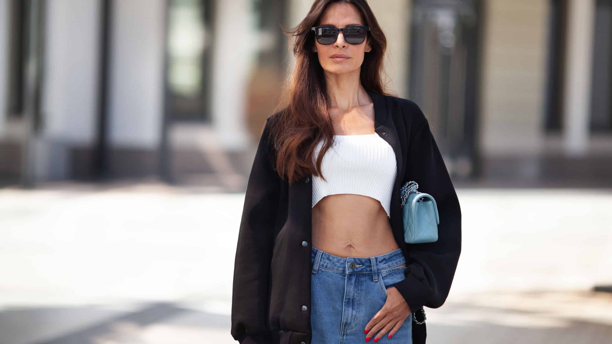 Portrait of beautiful fashionable brunette woman standing city street. Female model fashionista wears black bomber jacket, CROP TOP bare belly, jeans, sunglasses. Street style.