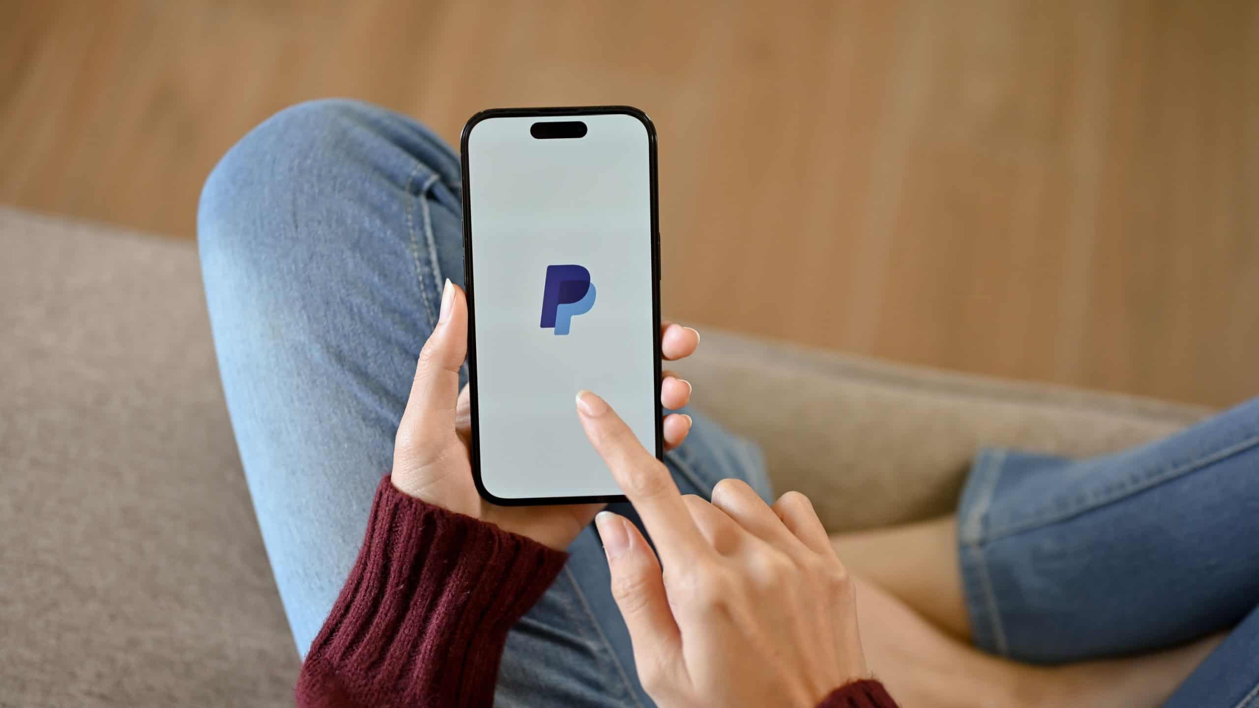 Chiang Mai, Thailand - July 25 2023: Top view of a woman in casual clothes using PayPal application on her smartphone while relaxing on a sofa in her living room. iPhone14 with PayPal logo on screen