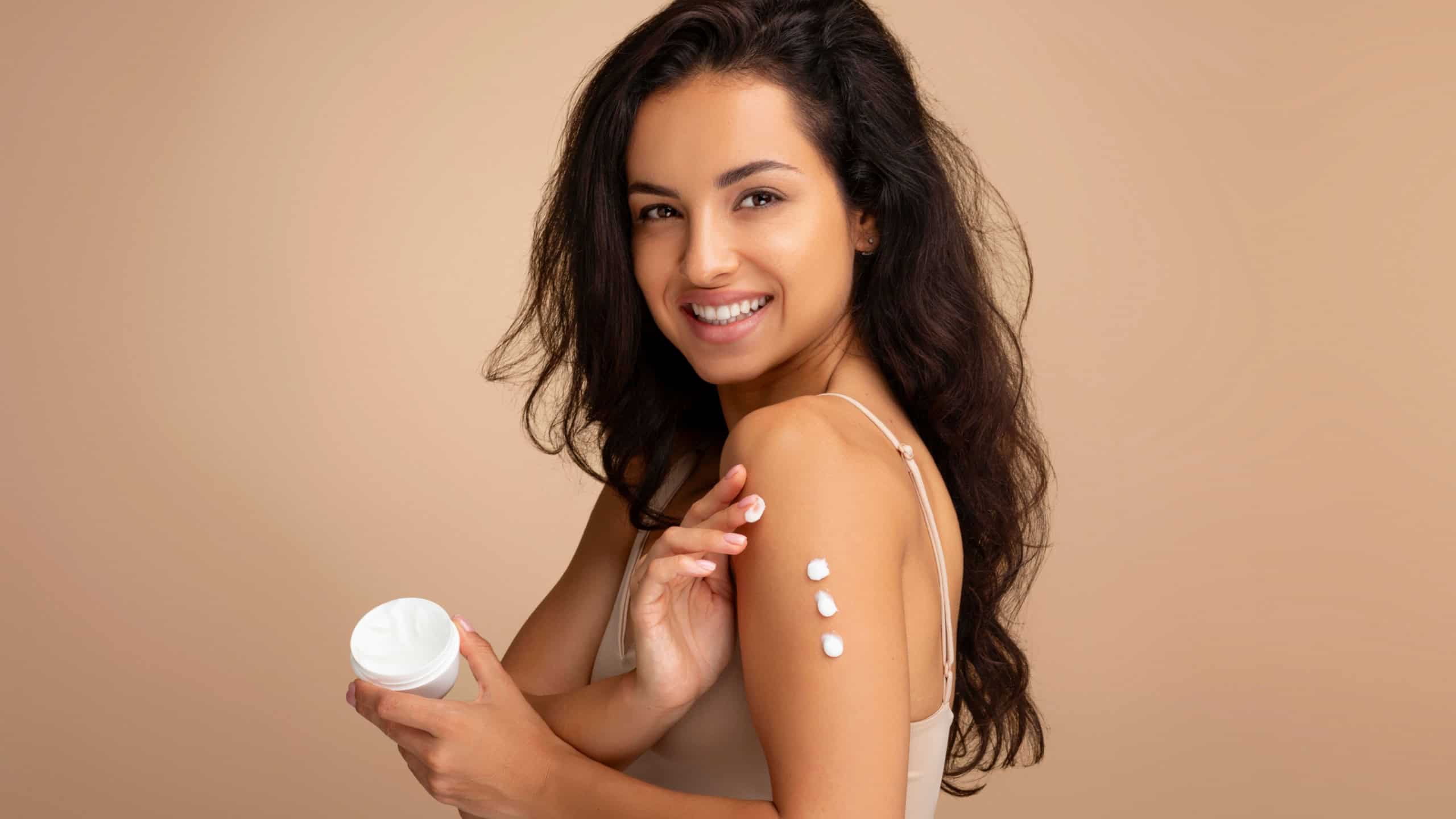 Happy attractive brunette middle eastern woman wearing beige top applying body cream on shoulder and smiling at camera, isolated on color background. Skin moisturizing, body care concept