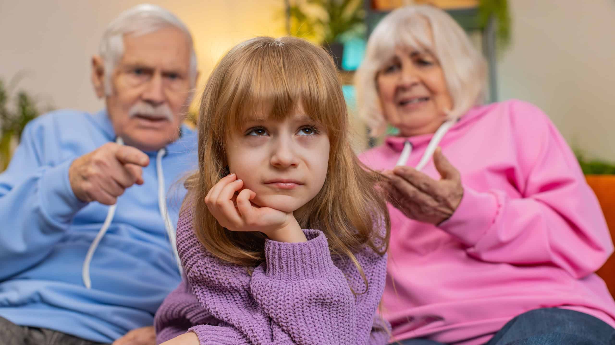 Irritated senior grandfather and grandmother scolding granddaughter for bad behavior and disobedience on sofa, raising voice, scream at little grandchild. Difficulties of upbringing, misbehaved child