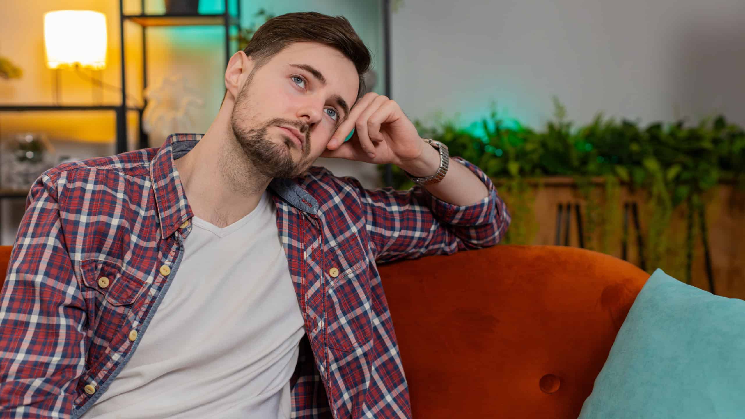 Disappointed young man sits at home reflecting on moral distress after quarrel break-up or divorce problem feeling bad annoyed ill sick. Sad Caucasian guy in room contemplates the emotional aftermath.