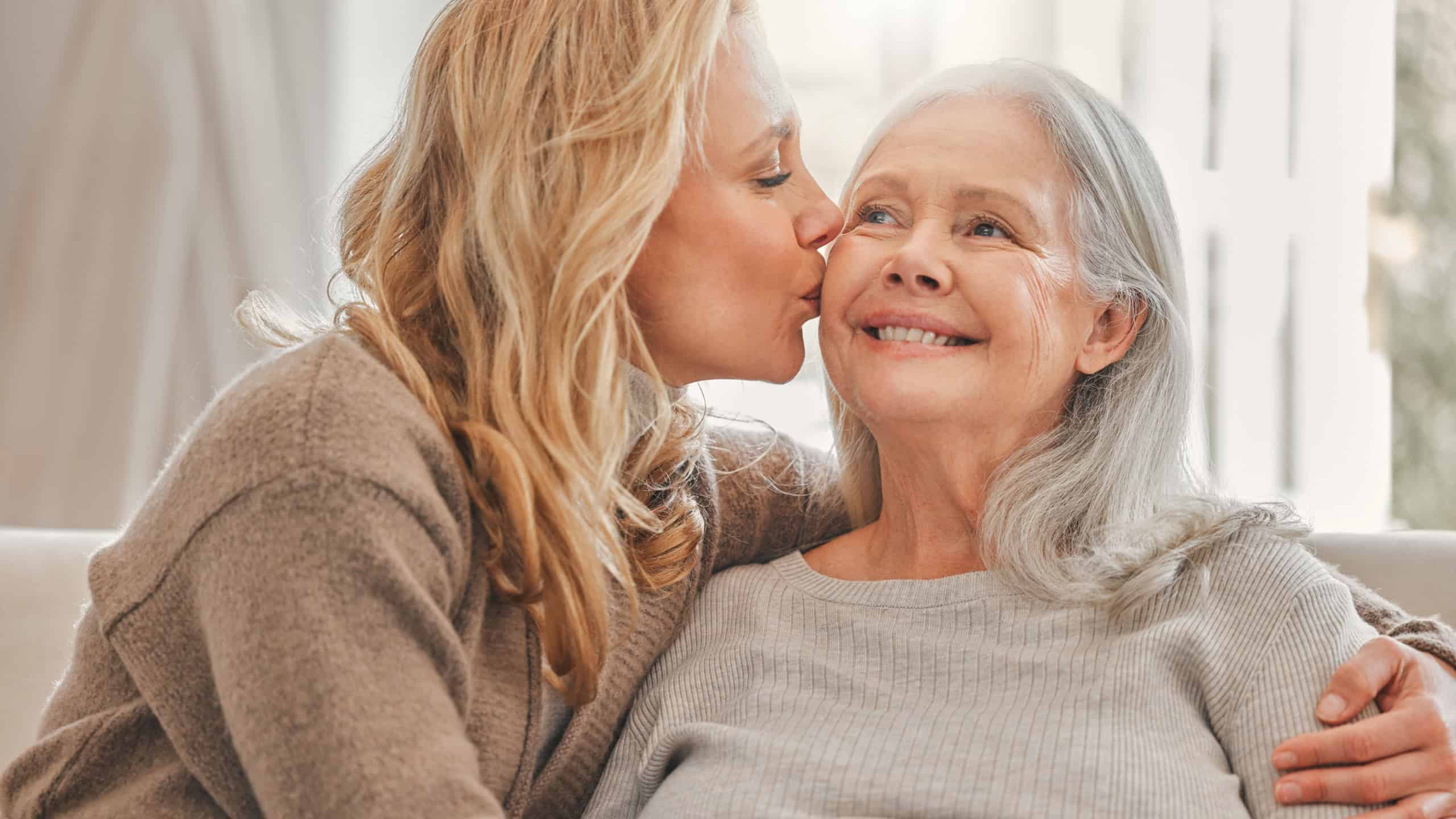 Hug, kiss or smile with senior mother and daughter on sofa in living room of home together for visit. Support, womens day or wellness with old parent and person embracing in apartment for bonding