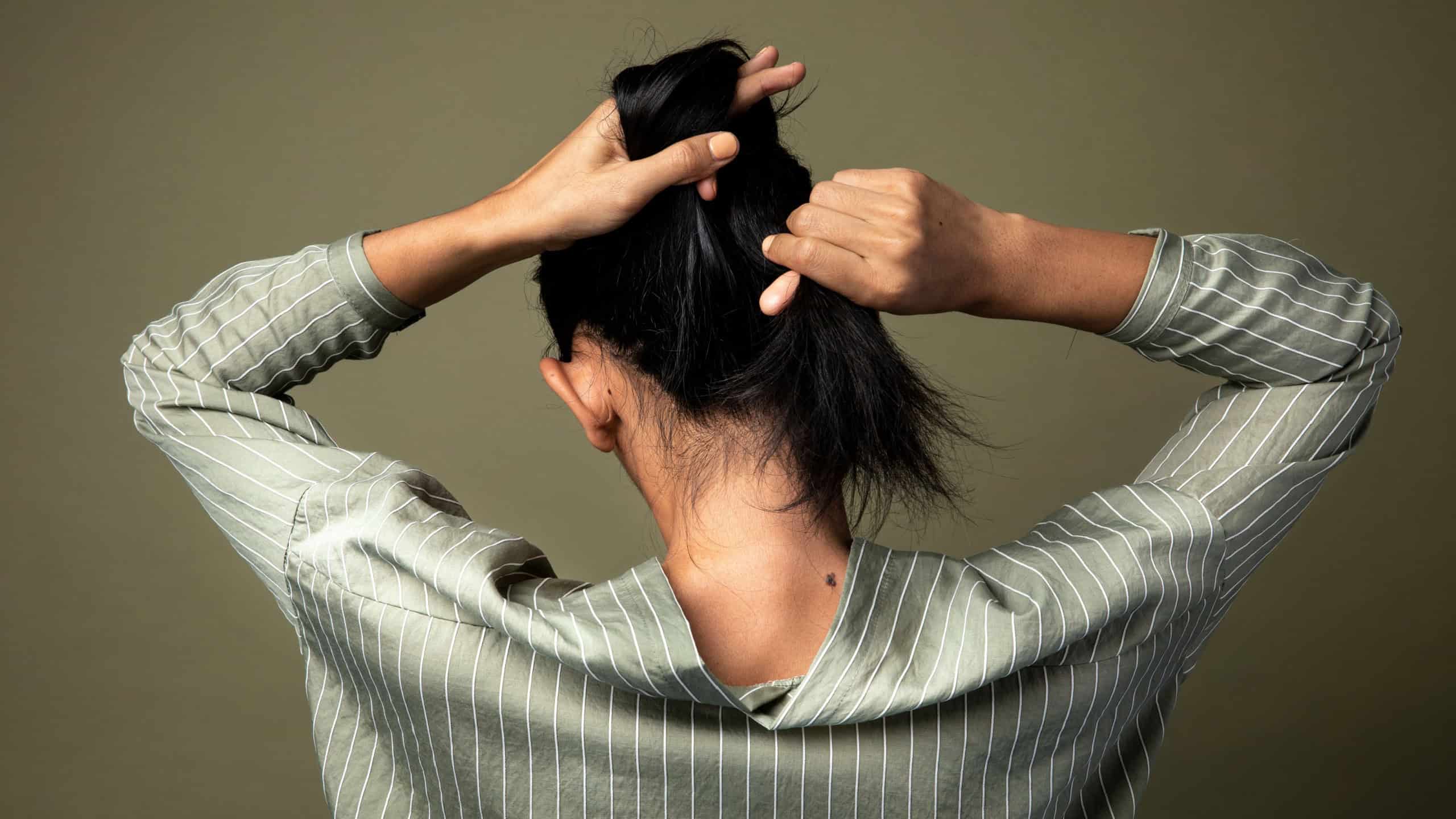 Asian woman tying her hair, back view. The woman is wearing a striped shirt. The woman is tying her hair into a bun. The woman is standing against green background. 