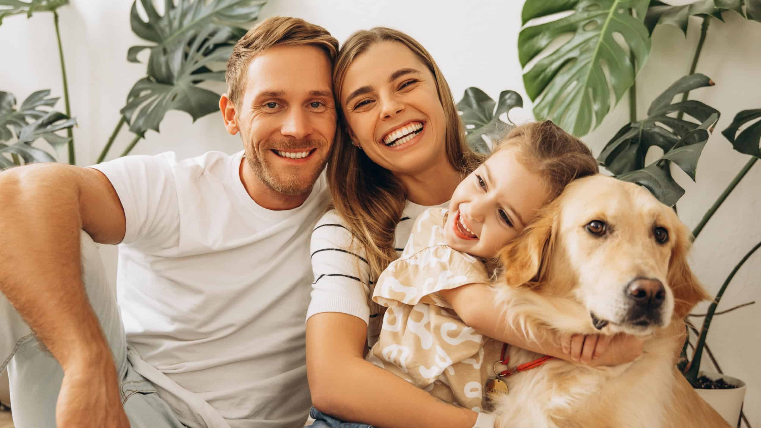 Portrait of attractive family, happy mother, father, cute, little daughter hugging together with dog, golden retriever at home, in cozy living room. Love, childhood concept