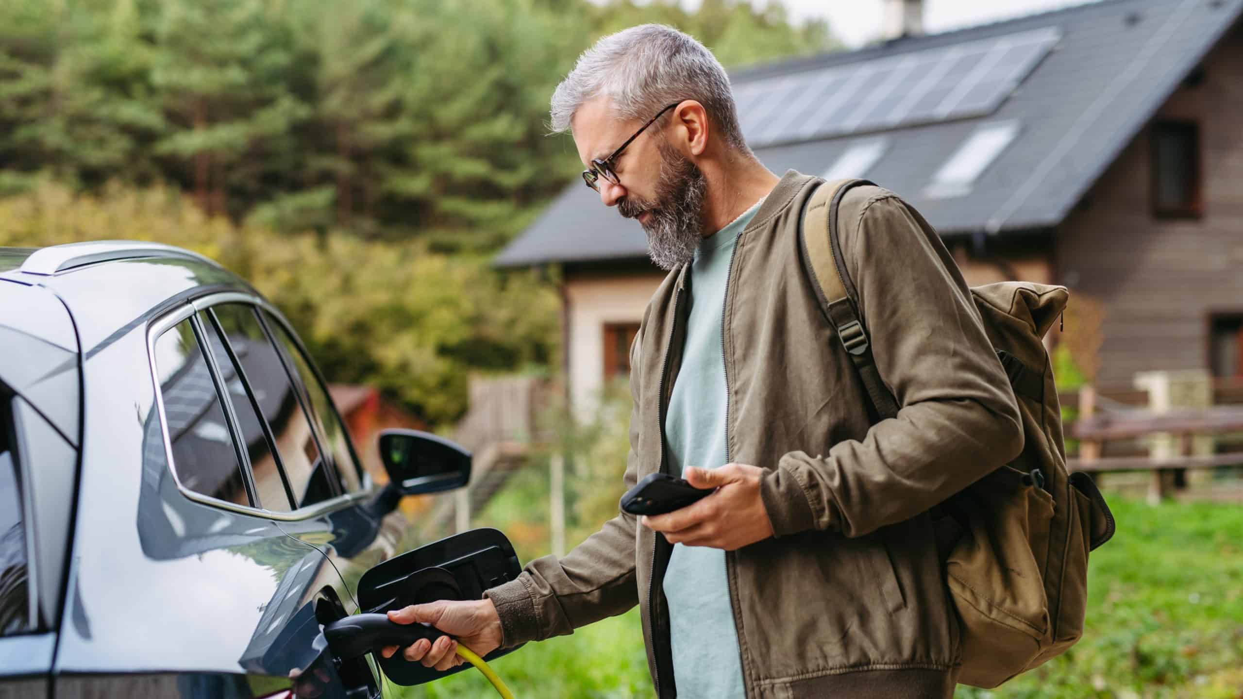 Man charging electric car in front of his house, plugging the charger into the charging port. House with solar panel system on roof behind him.