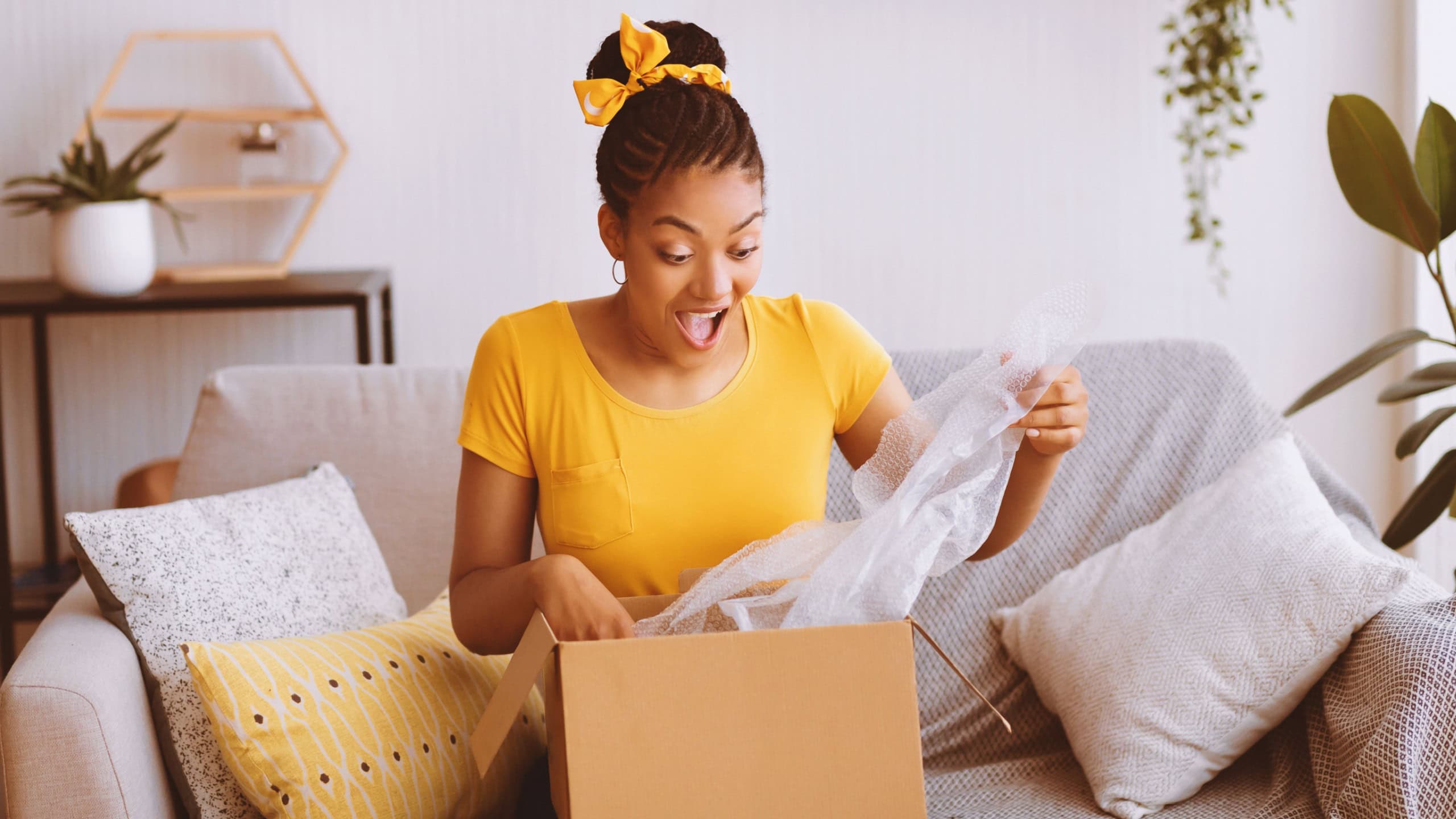 Fast Delivery. Overjoyed black woman received package, unpacking cardboard box, sitting on the sofa in living room at home, free space. Female buyer excited about her online shopping purchase