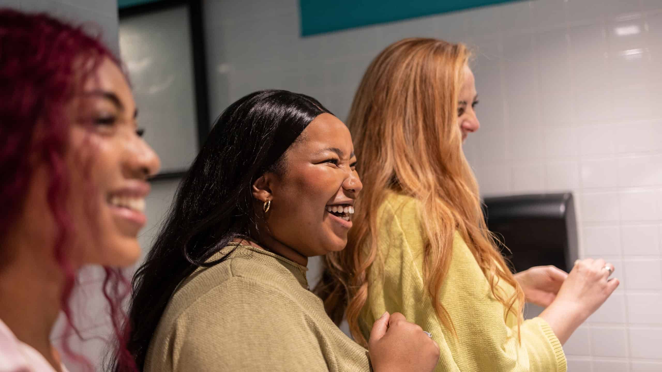Three diverse young women are enjoying a lighthearted moment together, laughing and talking in a public restroom, creating a sense of camaraderie and genuine connection
