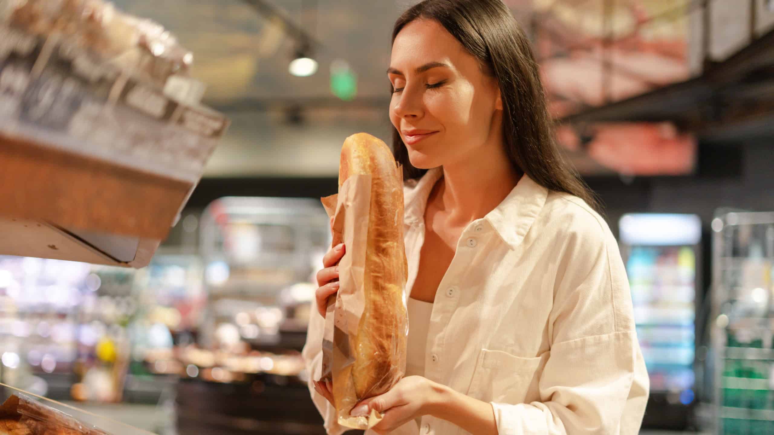 Beautiful smiling Latin woman enjoying the aroma of a freshly baked baguette while shopping in a supermarket bakery, surrounded by various bread and pastry products. Food concept