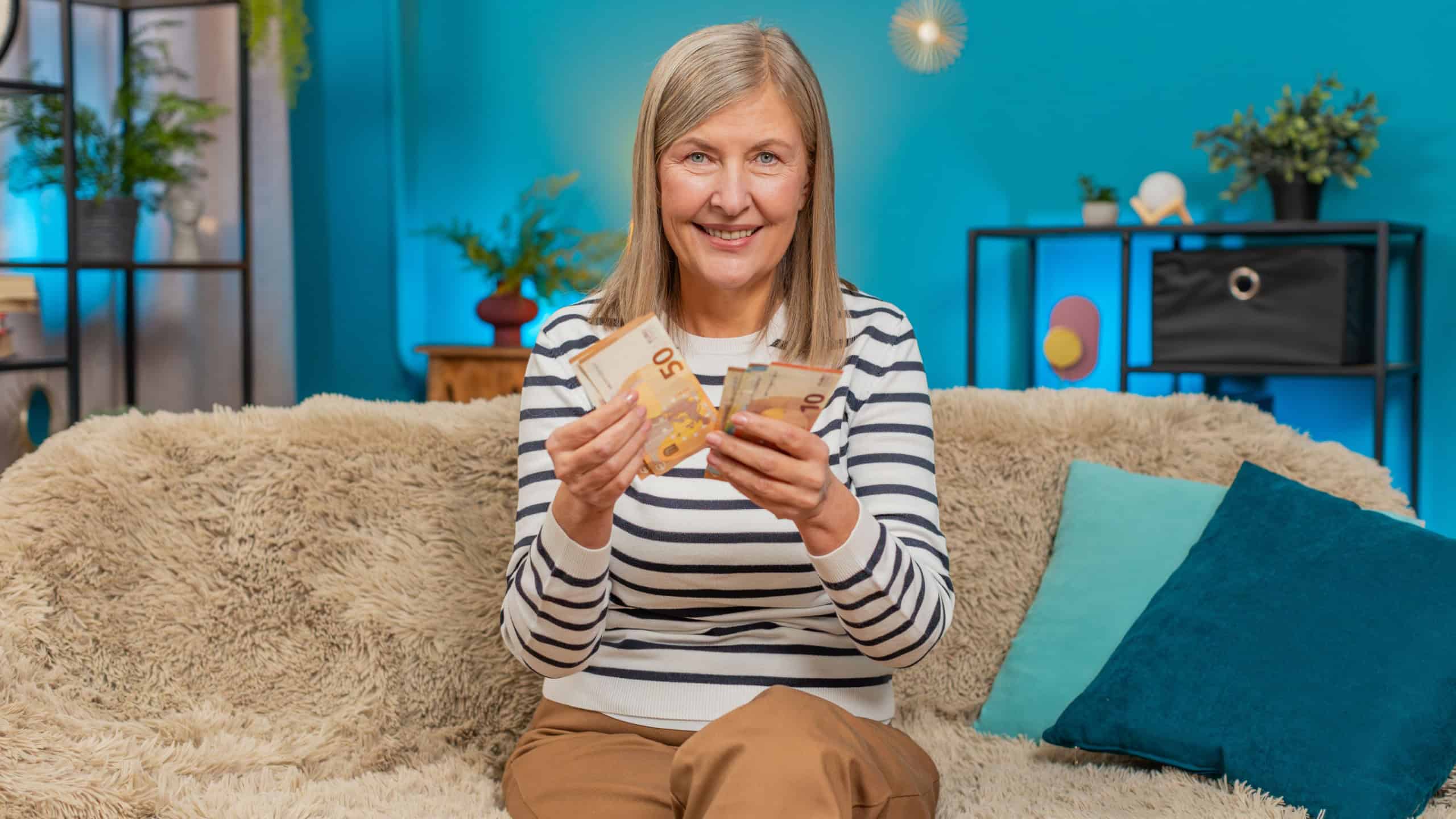 Happy Caucasian mature woman counts money euro cash in hands after winning lottery bonus or receiving a gift. Grandmother smiles broadly sitting on home sofa, celebrating financial victory salary