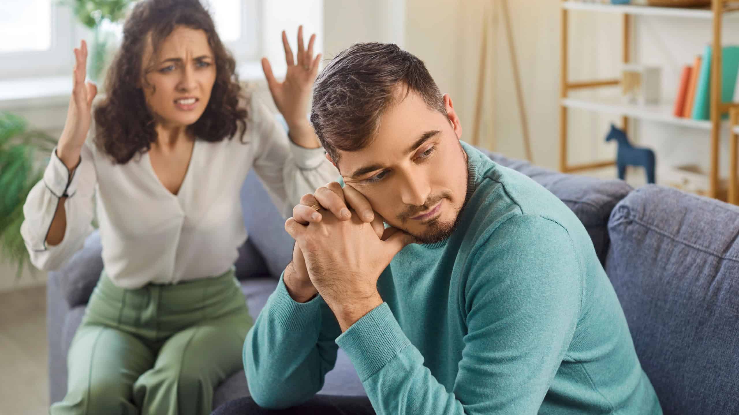 Guilty sad young man sitting on sofa, avoiding eye contact while angry upset woman shouting on him. Family couple arguing at home, having relationship problems, expressing anger and disappointment.