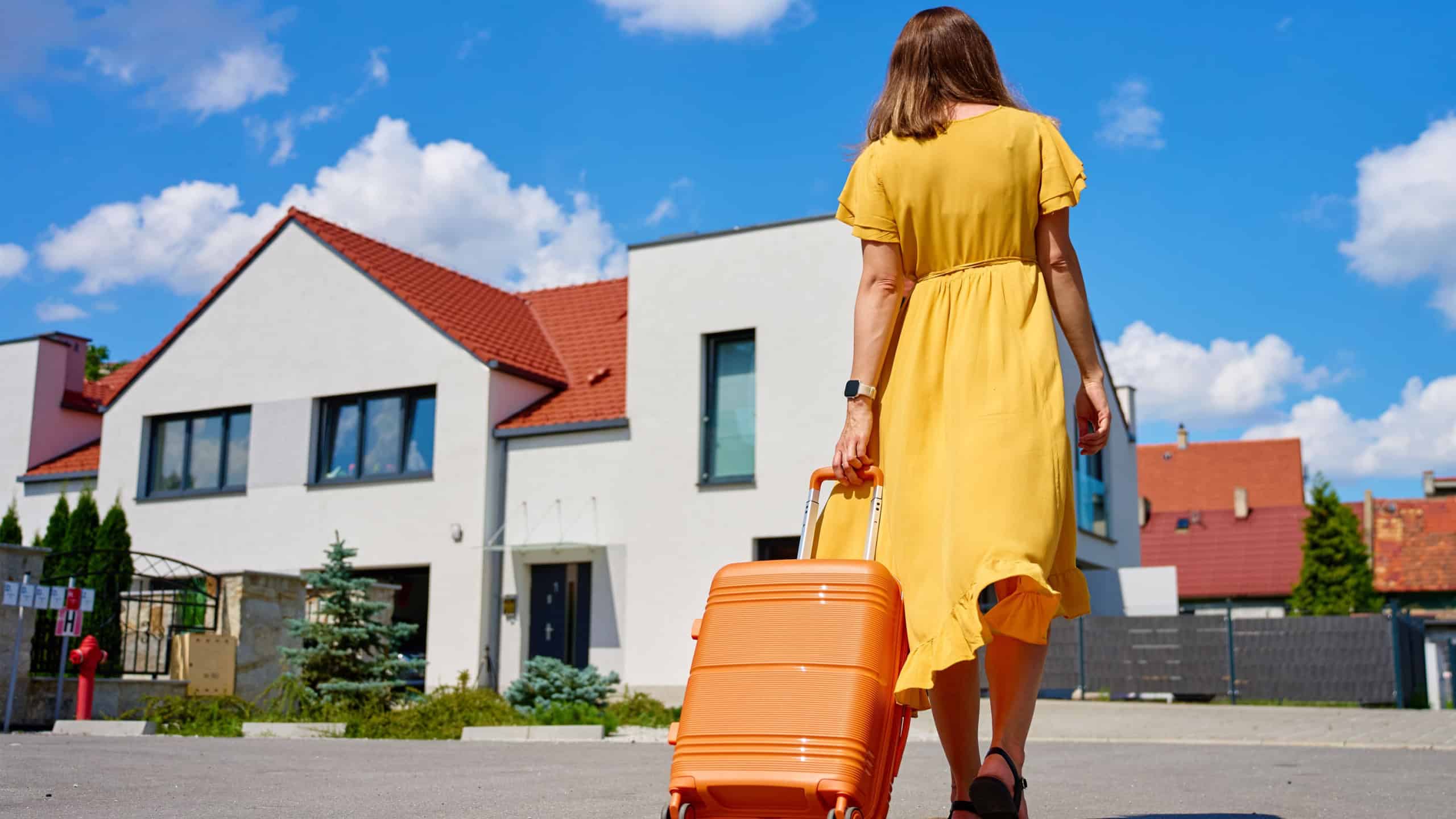 Woman in yellow dress pulls orange suitcase along residential street with houses on sunny day. Female traveler returning home from journey. Concept of travel, homecoming and end of vacation