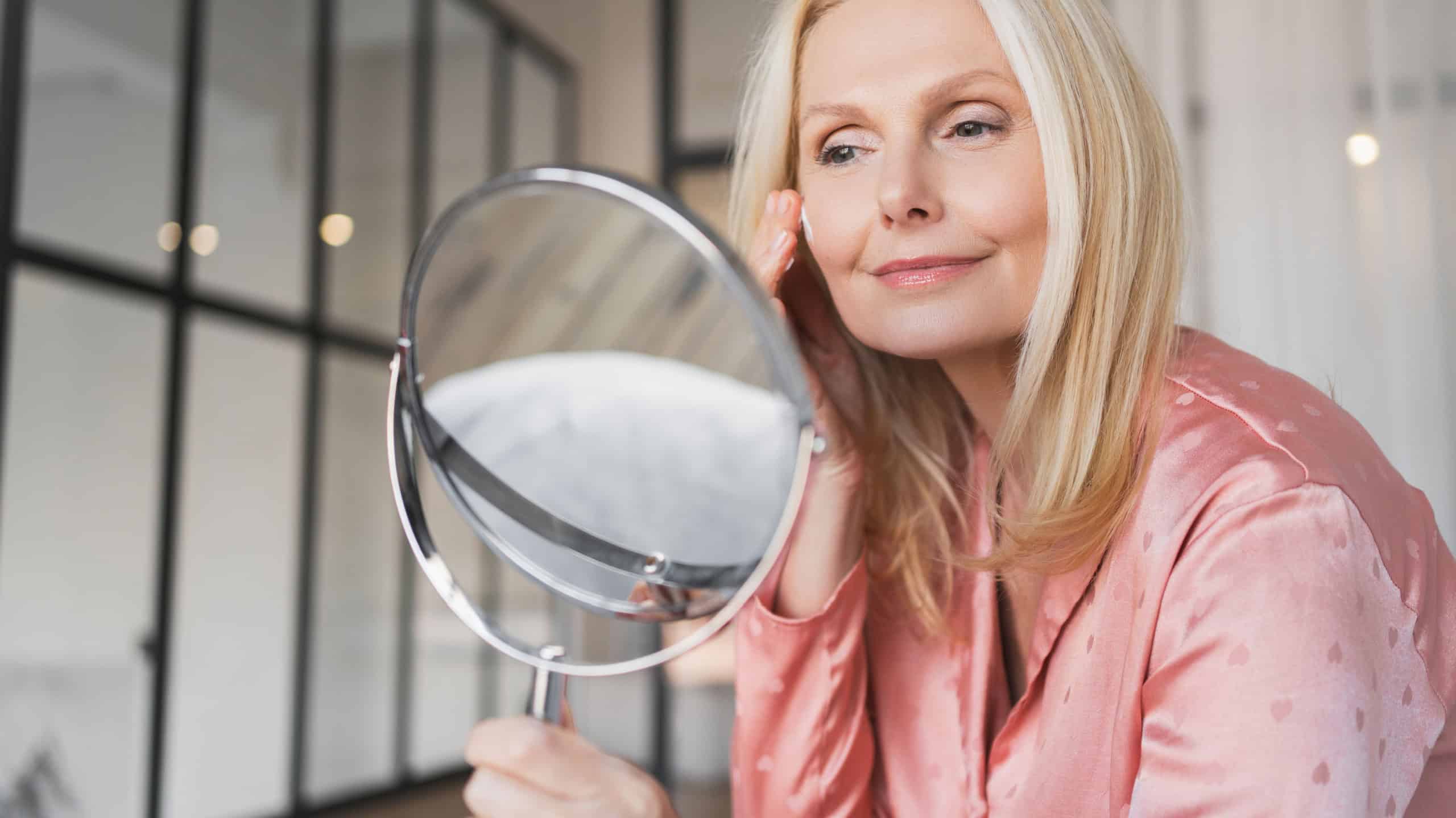 Close up of middle age Caucasian woman in sleepwear holding mirror using face cream at home, beautiful lady test newest organic beauty product, sitting on bed in light bedroom. Skin care routine.