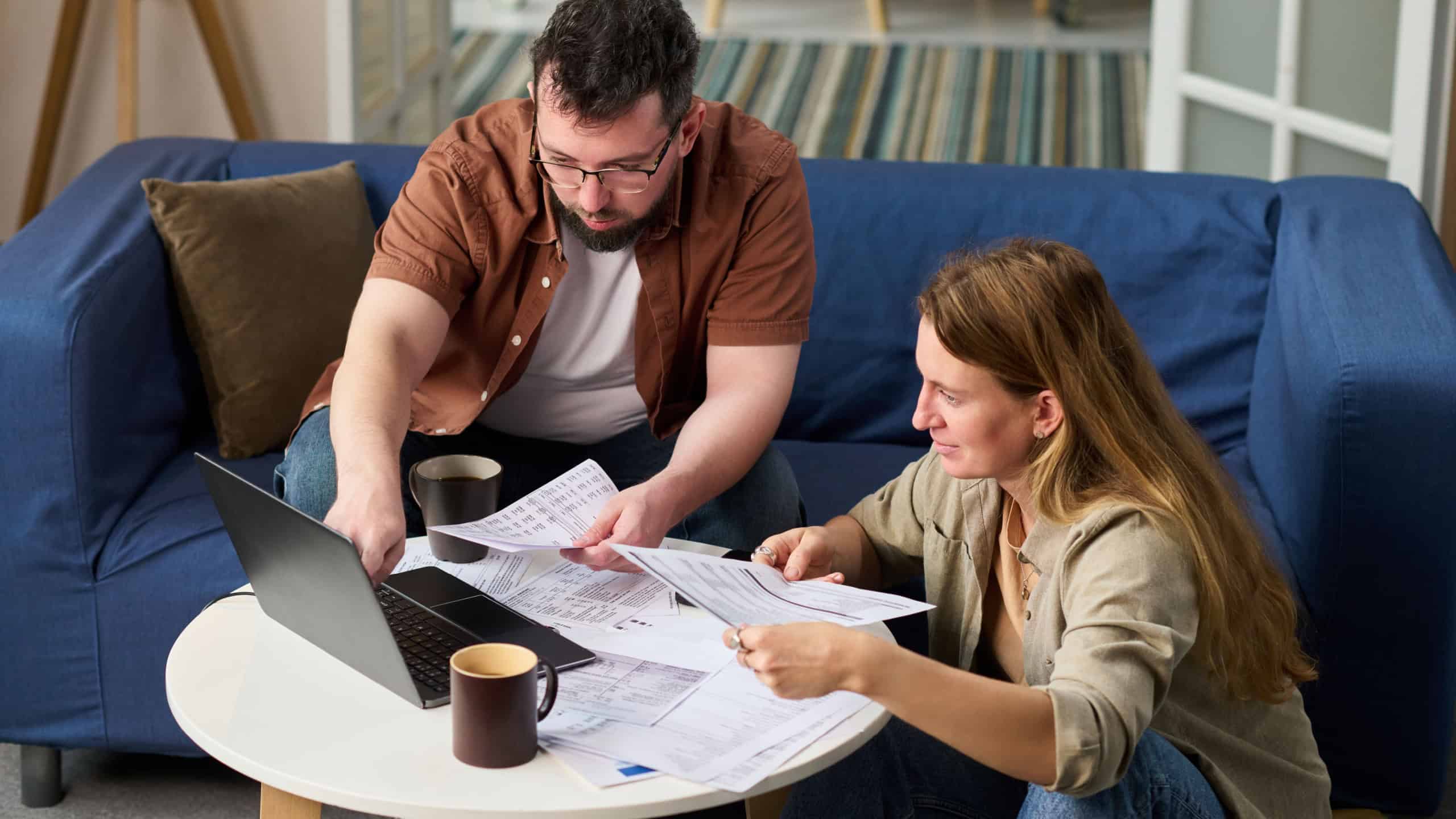 Caucasian middle aged man and Caucasian young adult woman sitting at table reviewing tax documents together using laptop, both focused on paperwork related to paying taxes